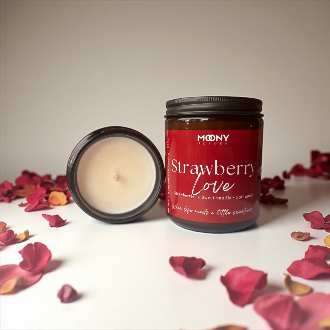 Moony Flames doftljus strawberry love i amber glasburk med röd etikett med vit text. Det syns några rosblad runt ljuset. Ljuset står framför en bejge bakgrund. 
