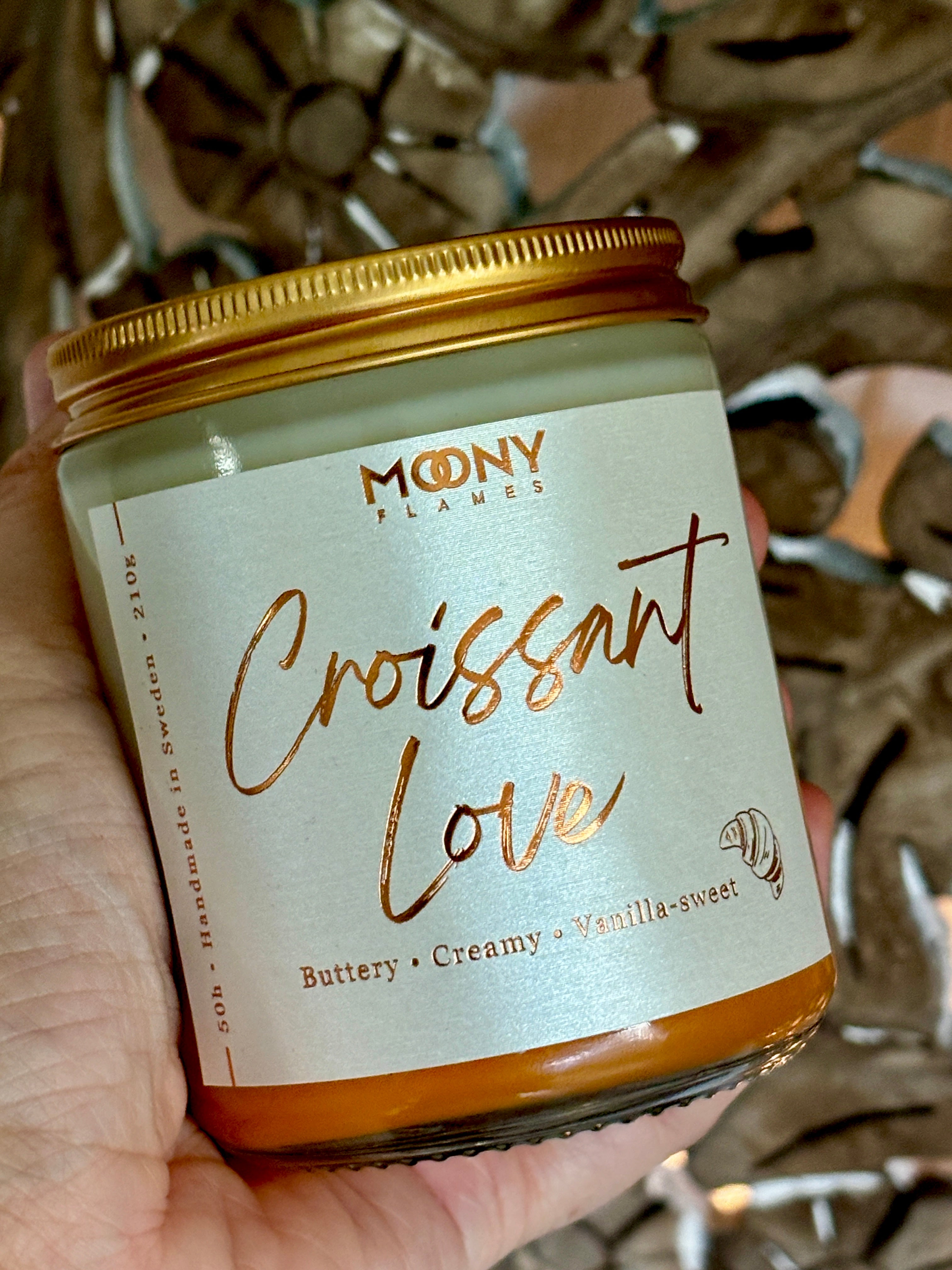 Doftljus Croissant Love i genomskinlig glasburk med skruvlock i guld som hålls i handen - Moony Flames.