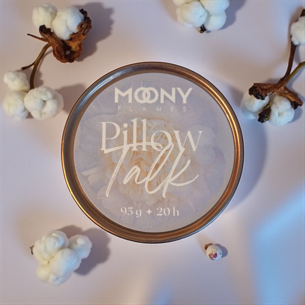 Bilden visar doftljuset Pillow Talk i metallburk i roséguld som står på en vit undergrund med bomull blommor runt sig.