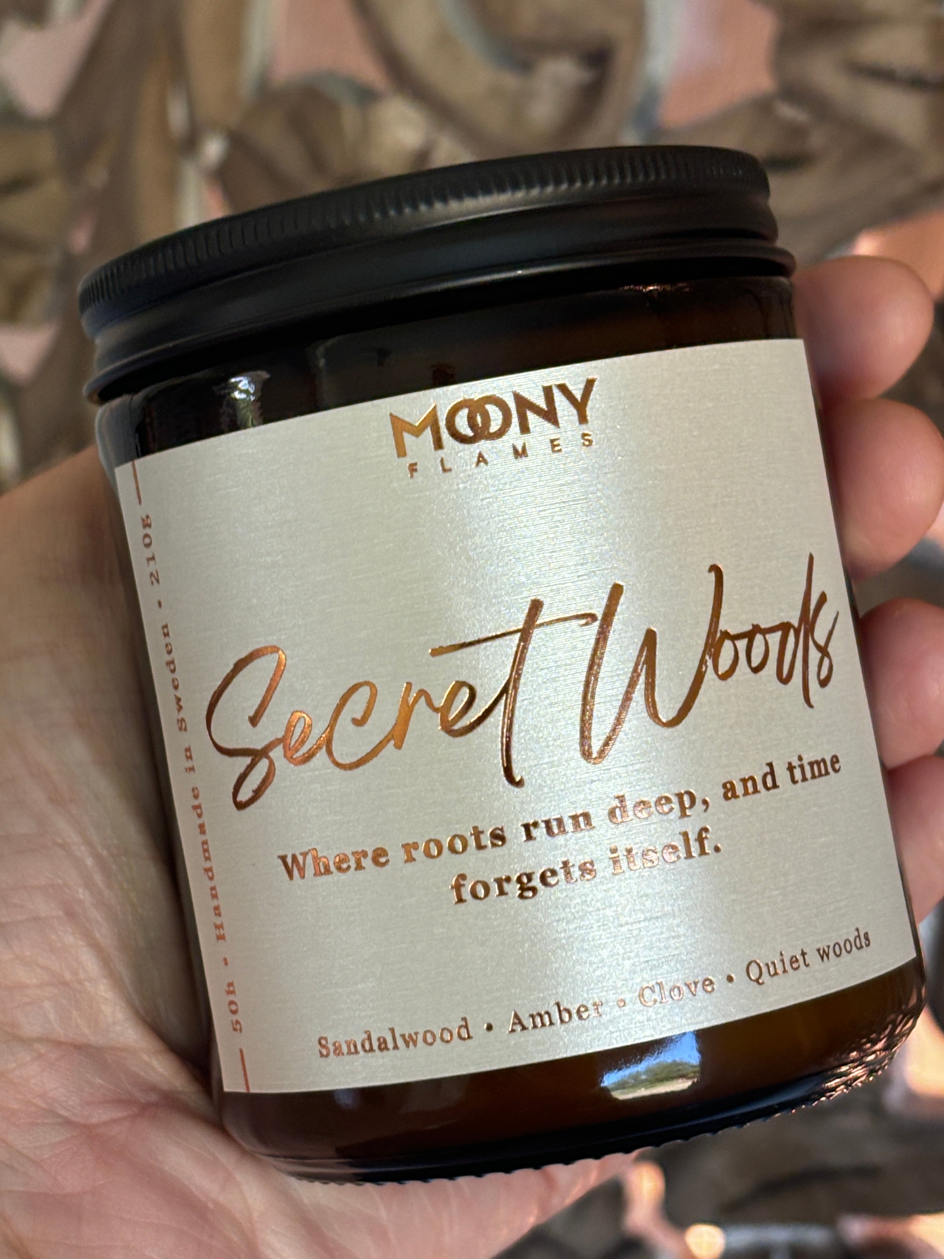 Doftljus Secret Woods i glasburk i amber med skimrande etikett och skruvlock i svart som hålls i handen  - Moony Flames
