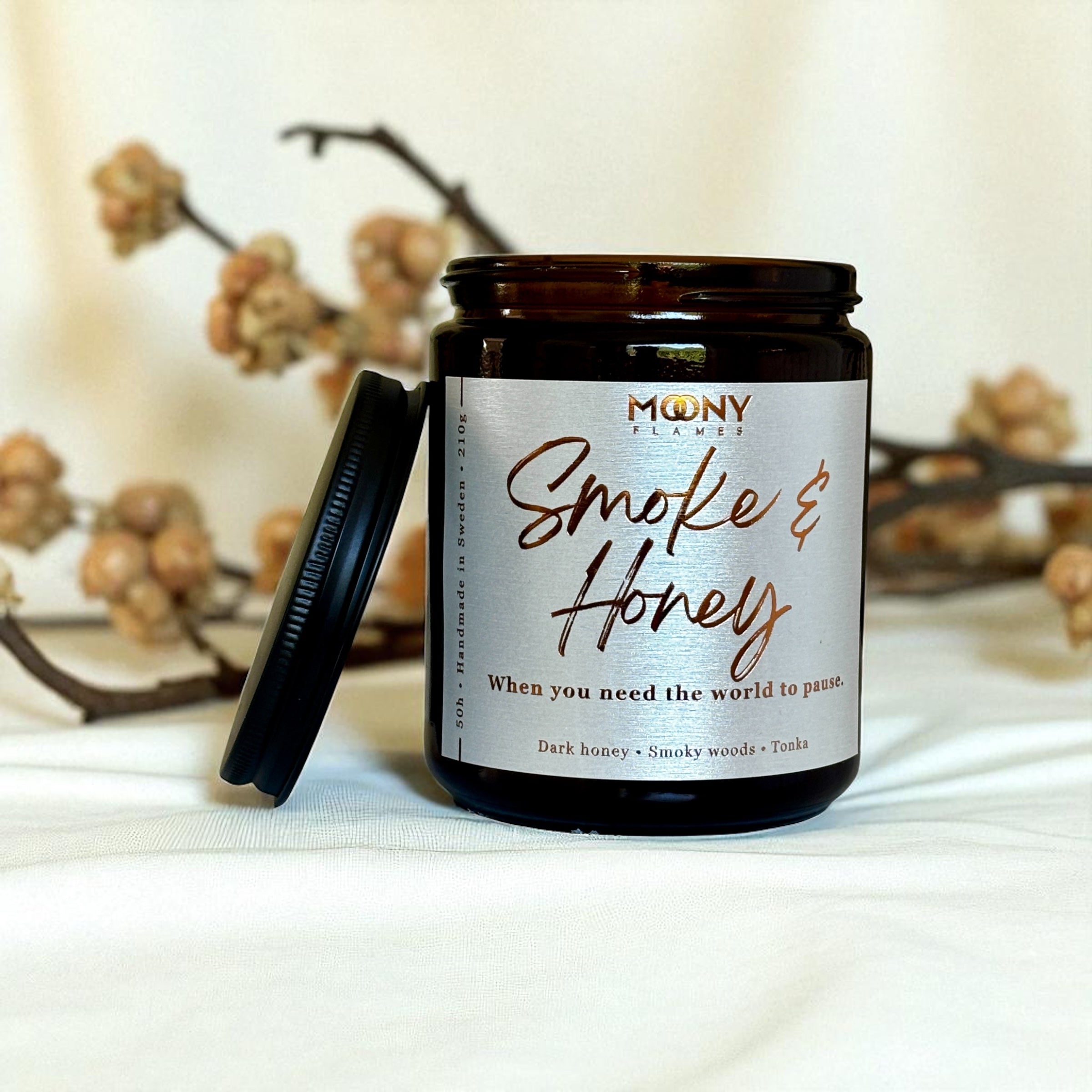 Doftljus Smoke and Honey i glasburk i amber med skimrande etikett och skruvlock i svart på en vit duk med torkade blommor i bakgrunden - Moony Flames