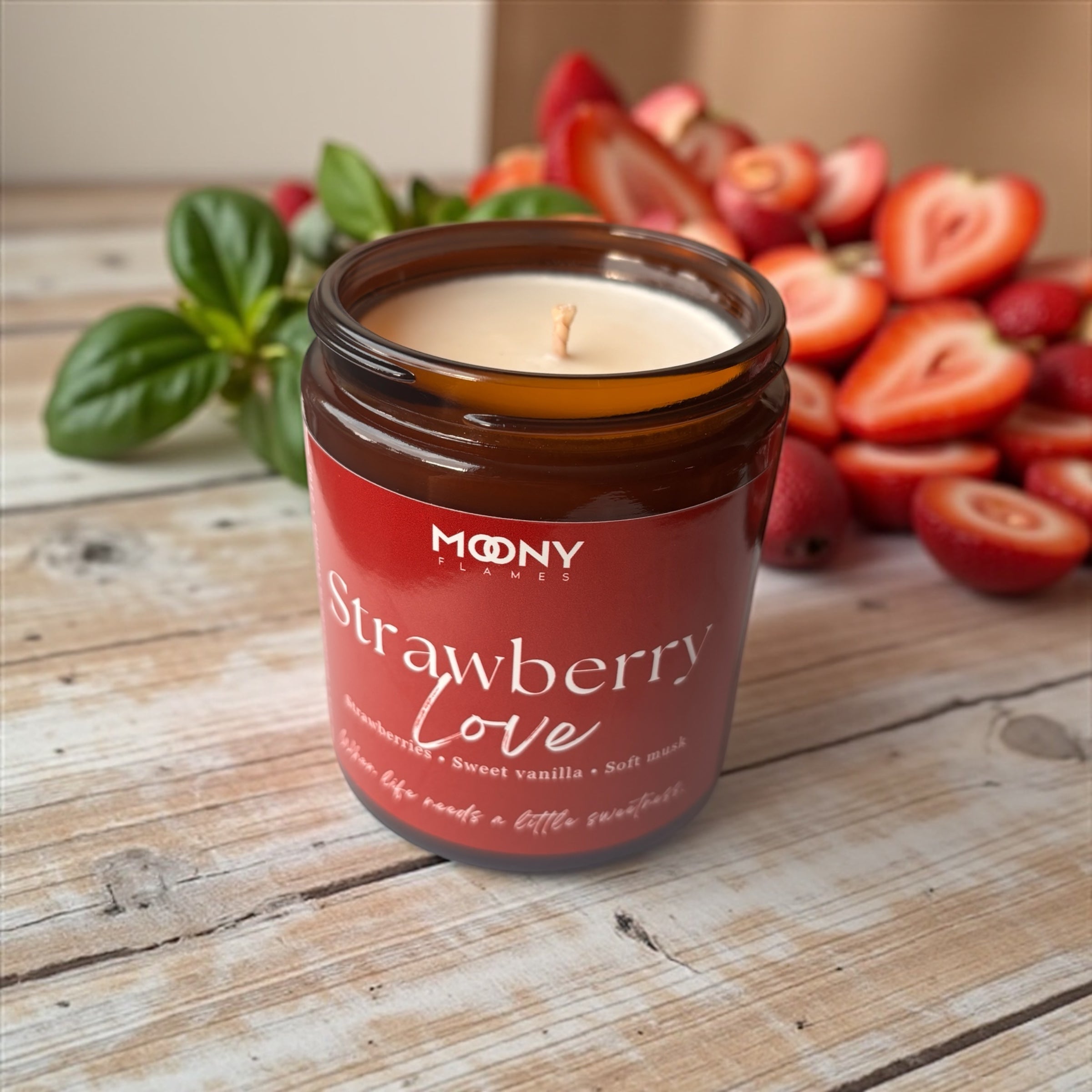 Bilden visar doftljuset Strawberry Love från Moony flames i glasburk i amber. Ljuset är öppet och står på ett rustikt bord  och det syns många jordgubbar och lite färskt basilika i bakgrunden.