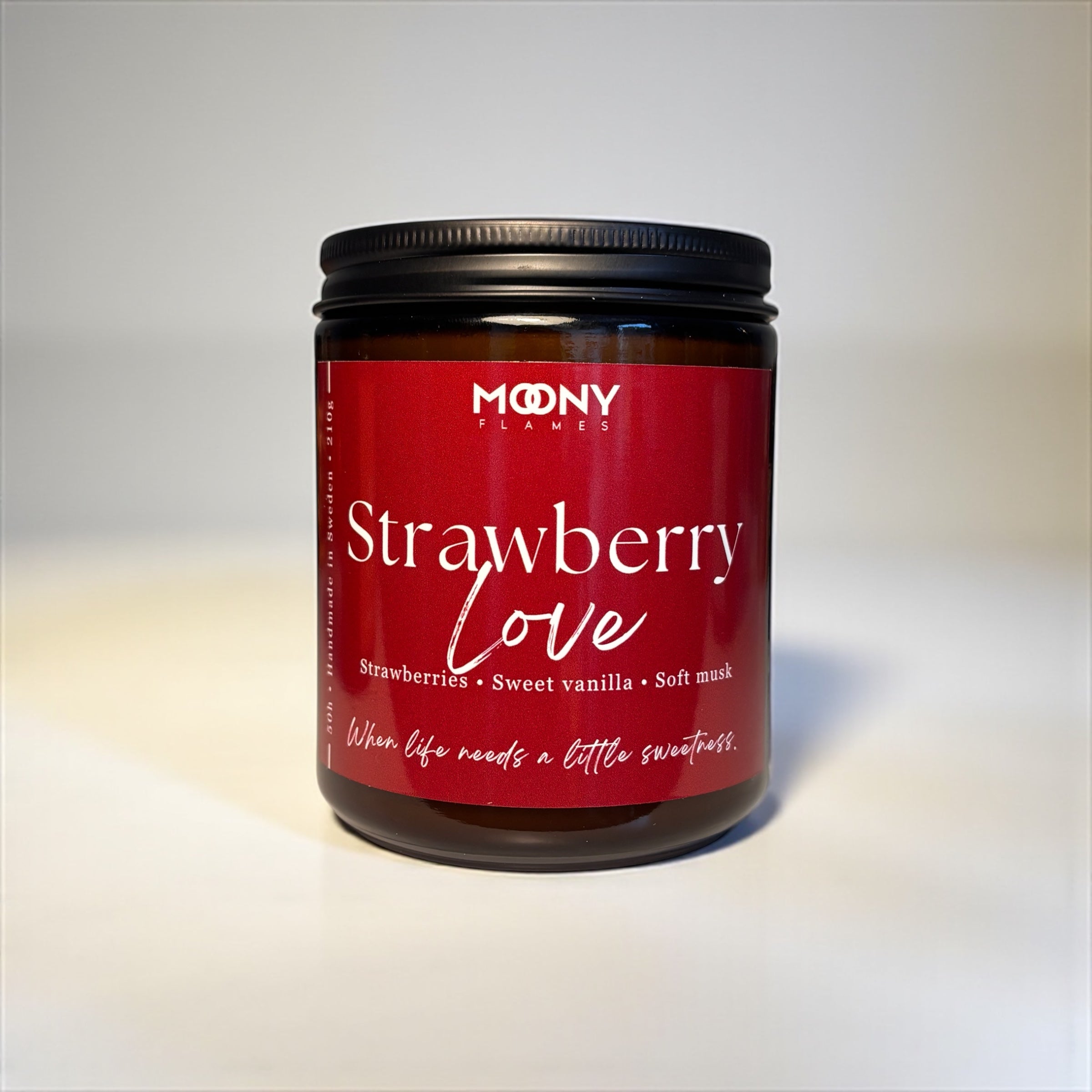 Doftljuset Strawberry Love från Moony Flames i glasburk i amber. Ljuset har ett svart skirvock och står framför en vit bakgrund.