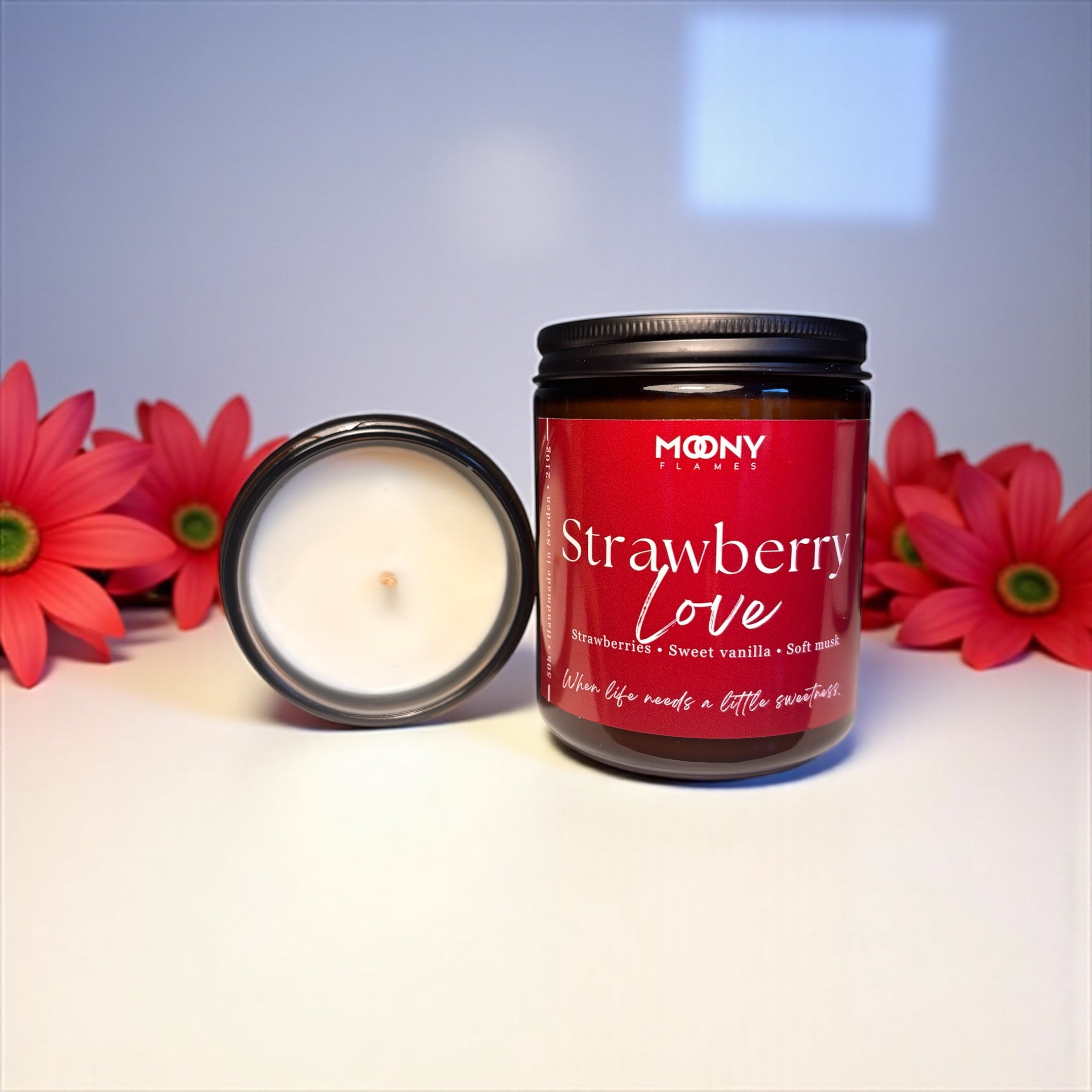 Bilden visar doftljuset Strawberry Love från Moony flames i glasburk i amber. Ljuset  står på en vit undergrund och det syns ett ljus liggandes bredvid, så man ser själva ljuset. Det finns röda blommor i bakgrunden.