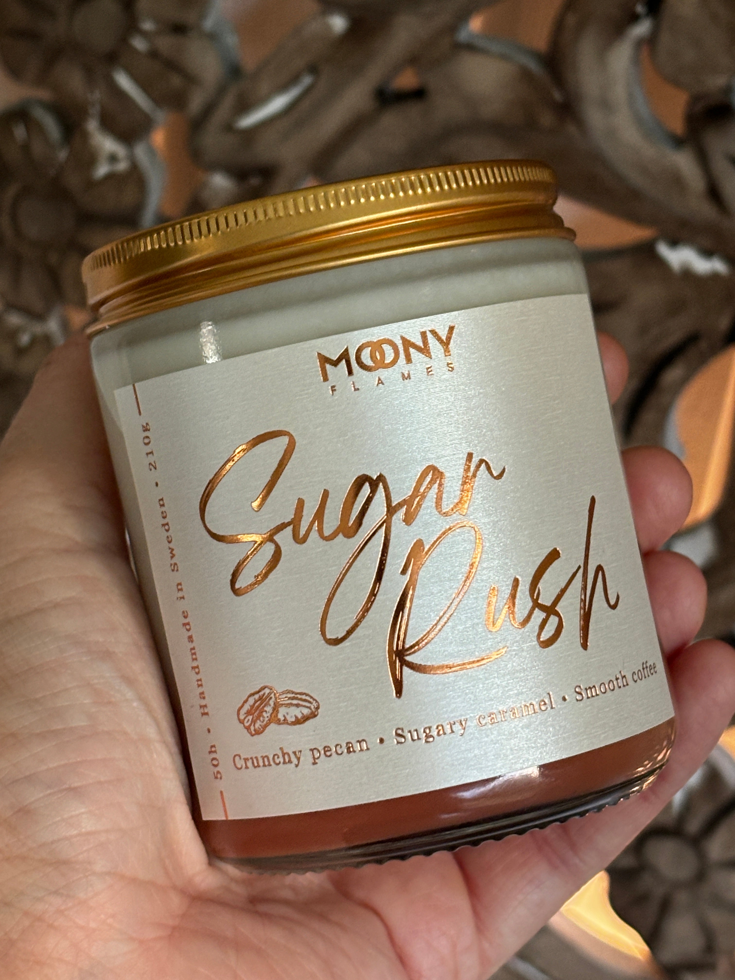 Doftljus Sugar Rush i genomskinlig glasburk i med skimrande etikett och skruvlock i guld som hålls i handen - Moony Flames