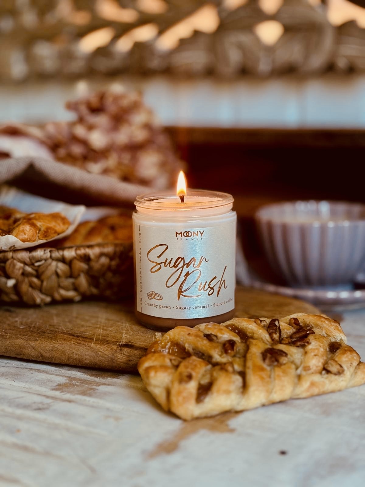 Doftljus Sugar Rush i en genomskinlig burk som står tänt på ett rustikt bord. Det syns pecan danish bakelser, en kaffekopp bredvid. Moony Flames
