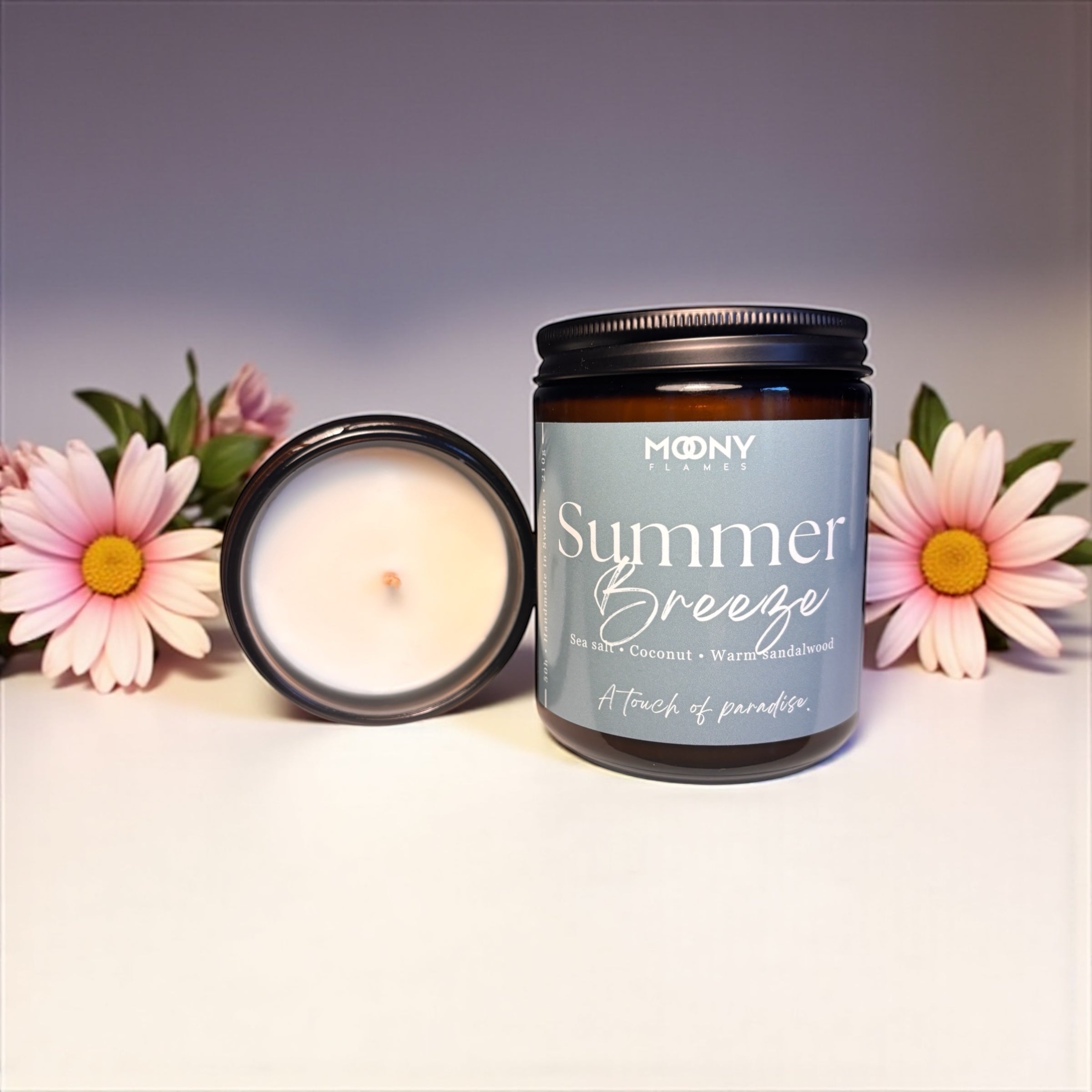 På bilden visas doftljuset Summer Breeze från Moony Flames ståendes på en vit bakgrund med somriga blommor i bakrunden. Glasburken i amberfärg har en snygg ljusblå etikett och visas framifrån. 