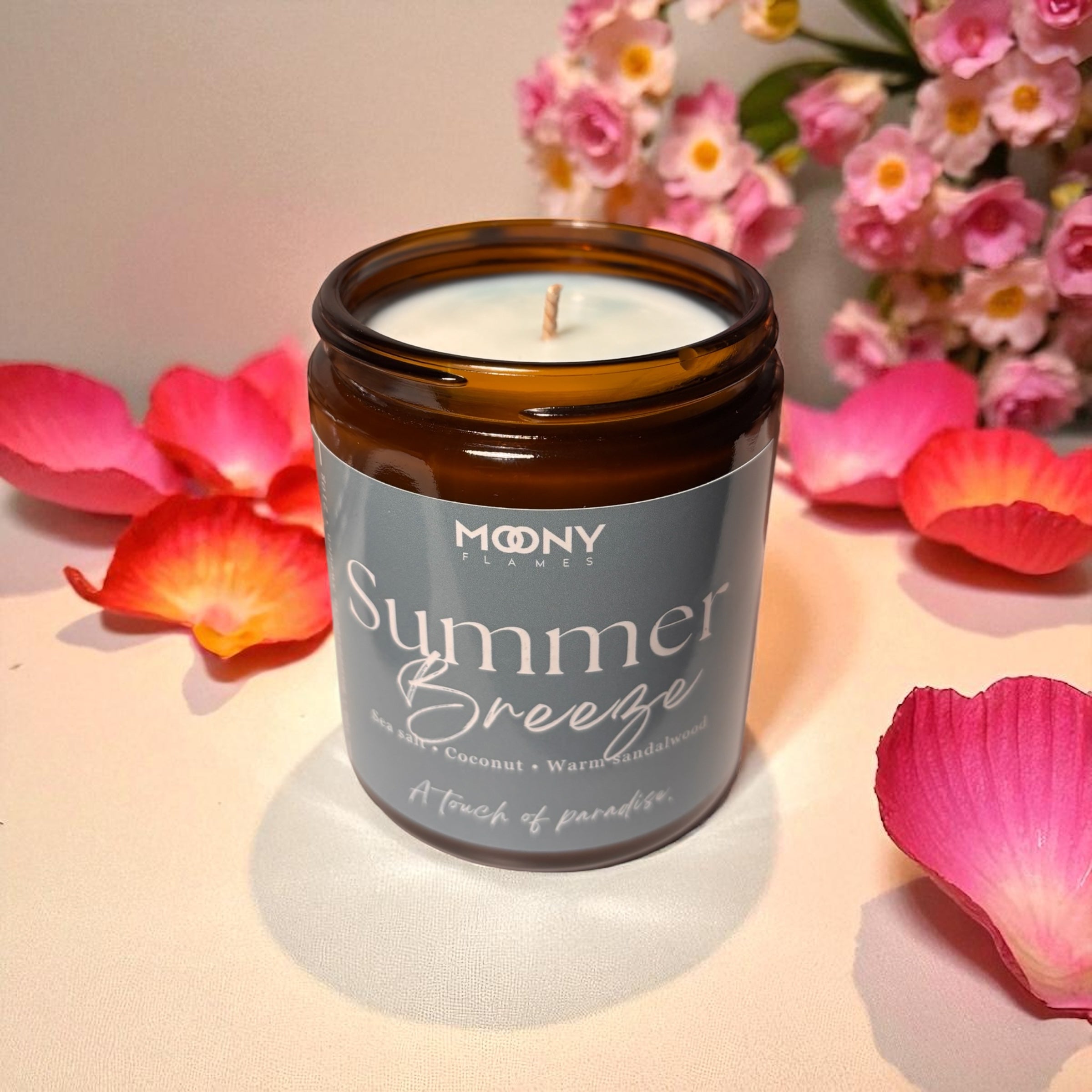 På bilden visas doftljuset Summer Breeze från Moony Flames ståendes på en vit bakgrund med somriga blommor i bakrunden. Glasburken i amberfärg är öppen har en snygg ljusblå etikett och visas framifrån. 
