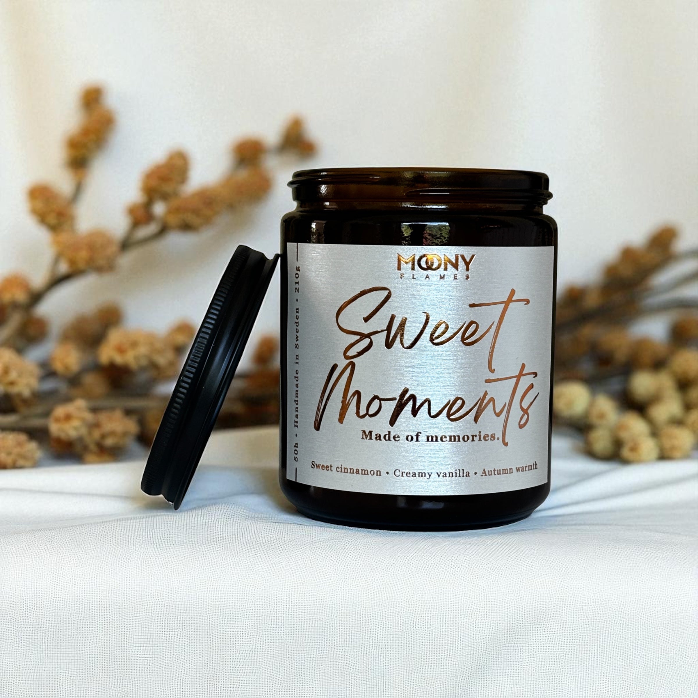 Doftljus Sweet Moments i glasburk i amber med skimrande etikett och svart lock. Ljuset står på en vit duk med torkade blommor i bakgrunden - Moony Flames.