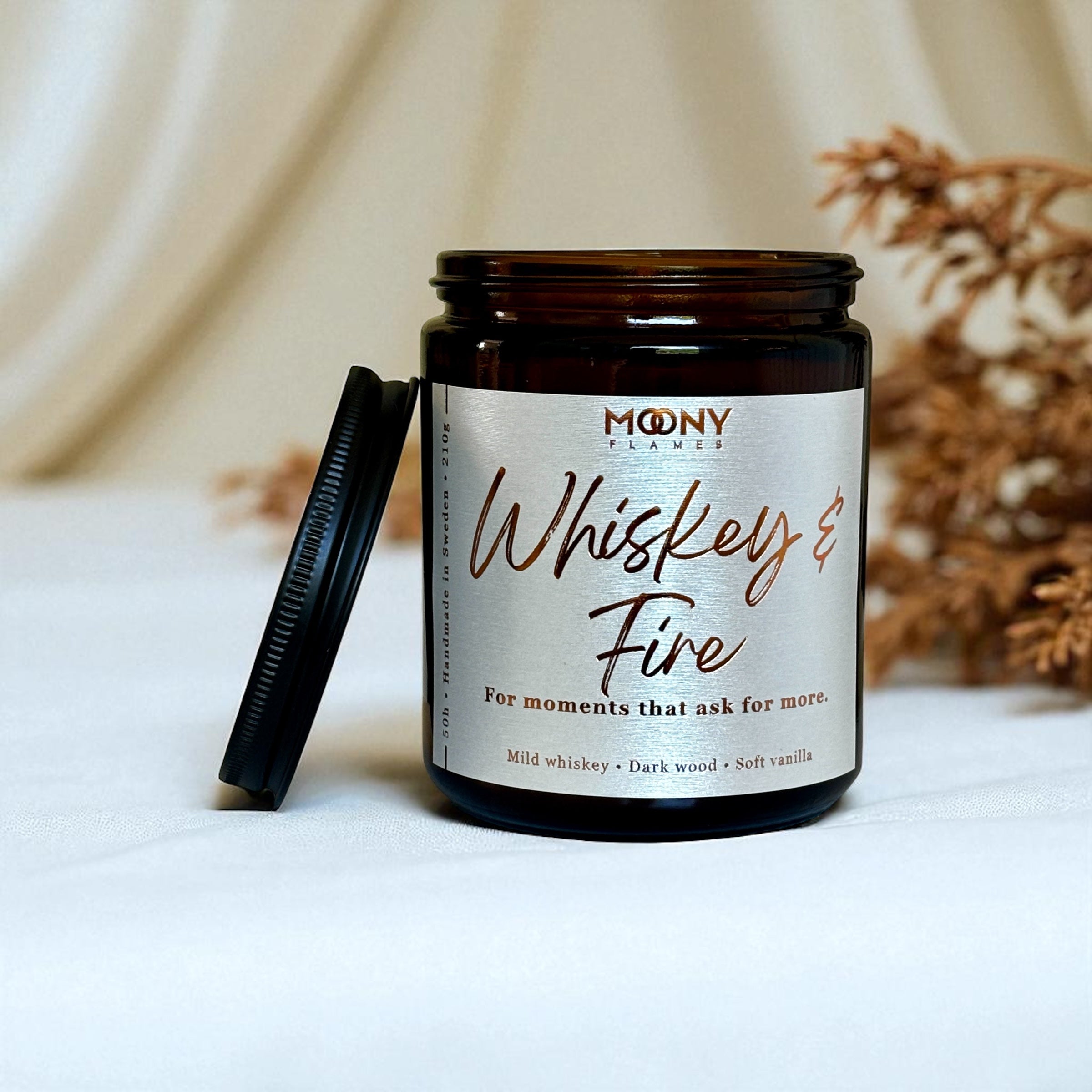 Doftljus Whiskey and Fire i glasburk i amber med skimrande etikett och svart lock. Ljuset står på en vit duk med torkade blommor i bakgrunden - Moony Flames.