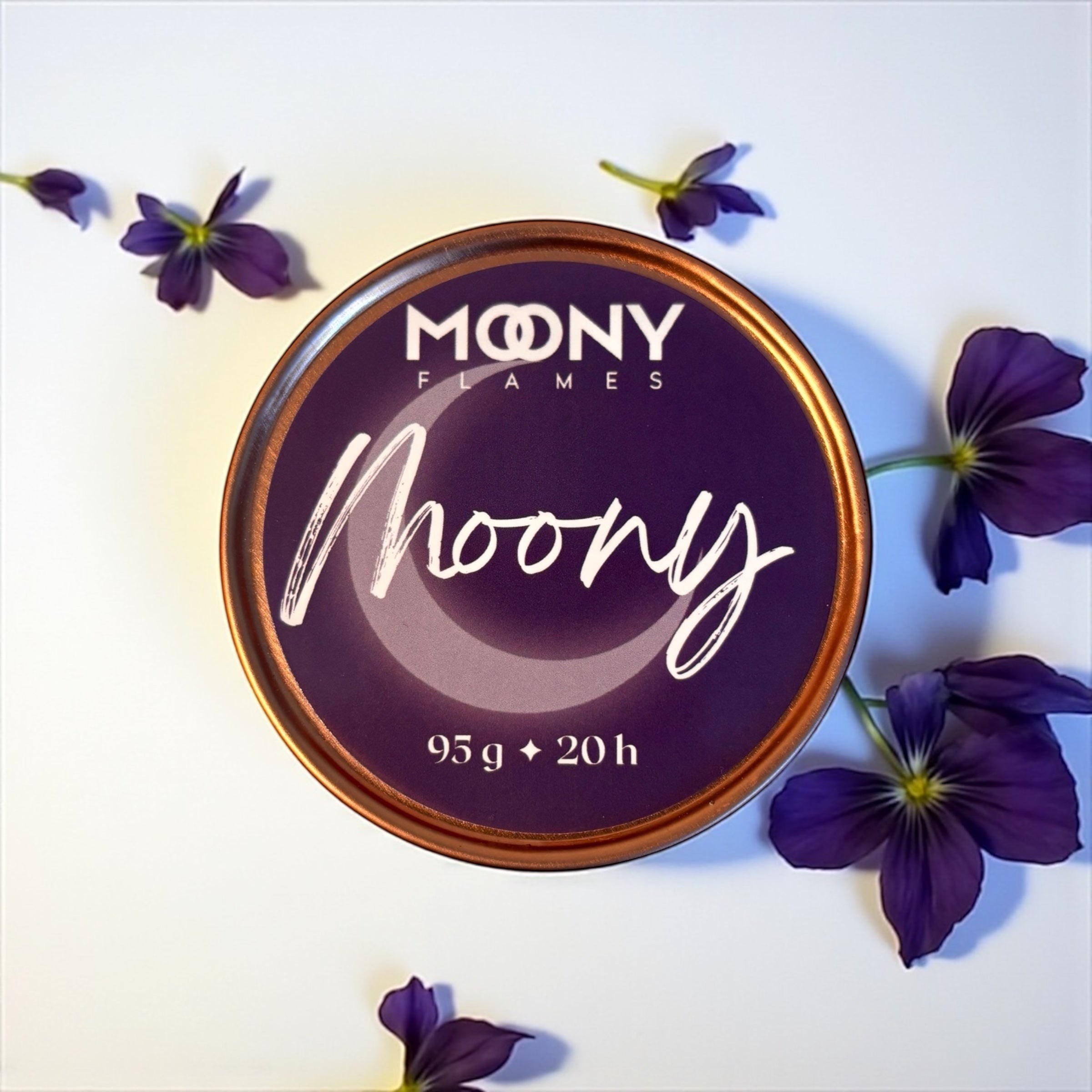 Bilden visar doftljuset Moony från Moony Flames i en metallburk i roséguld som står på en vit undergrund. Runt ljuset ligger mörklila blommor som dekoration.