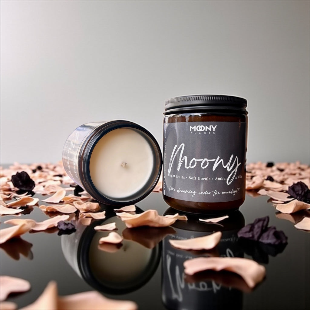 Bilden visar doftljuset Moony från Moony Flames som står på en svart speglande yta med många rosblad runt sig och ett liggnade ljus bredvid så man kan se själva ljuset i burken.