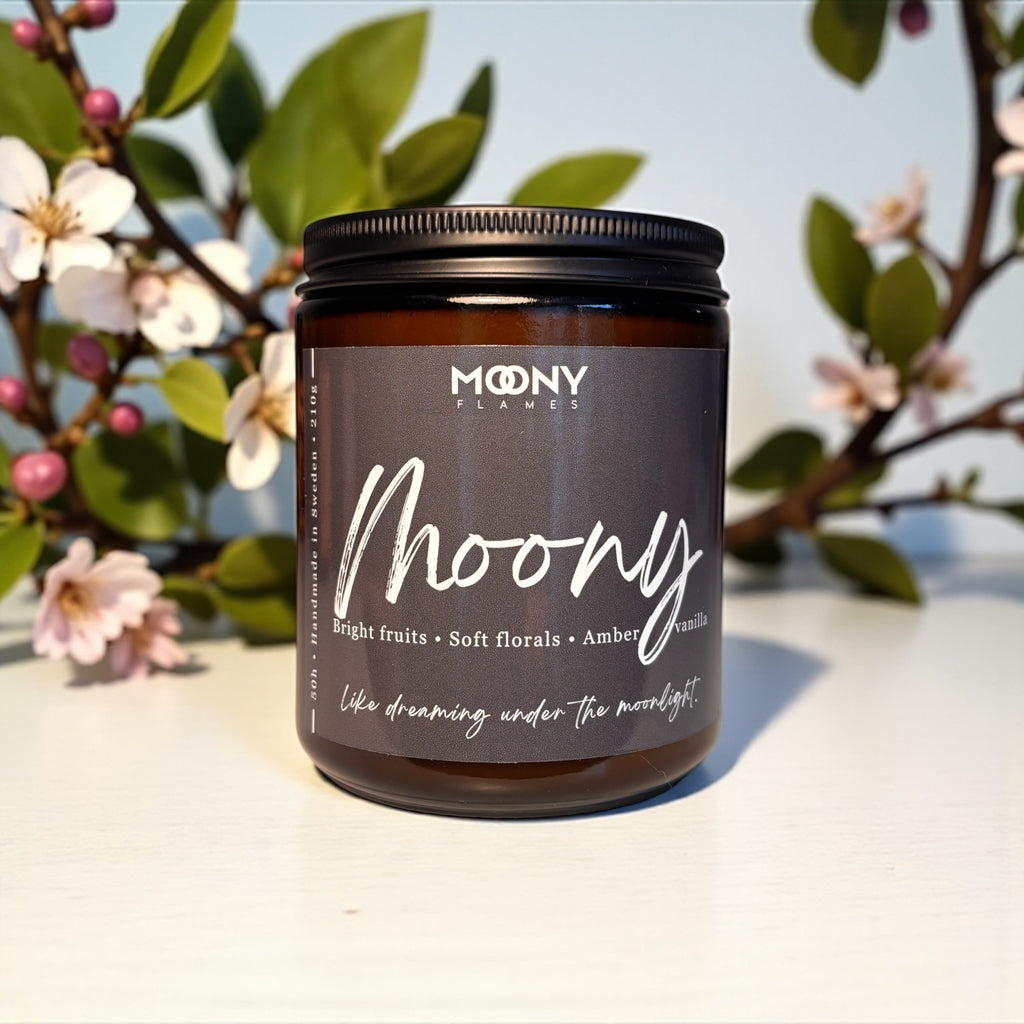 Här visas doftljuset Moony från Moony Flames i en amber glasburk med svart skruvlock. Ljuset står på en vit undergrund och det sisas vårblommor i bakgrunden. 