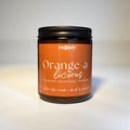 Här ser man doftljuset Orange-a-licious  i en glasburk i amber som är professionellt fotograferat framför en vit bakgrund.
