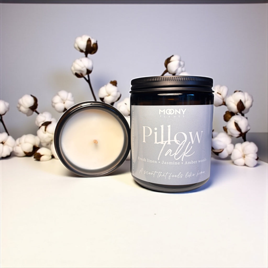 På bilden visas Moony Flames doftljus Pillow Talk i glasburk i amber som står framför en vit bakgrund. Det syns många bomulls blommor bakom ljuset. En öppnad burk ligger bredvid så man an se själva ljuset.