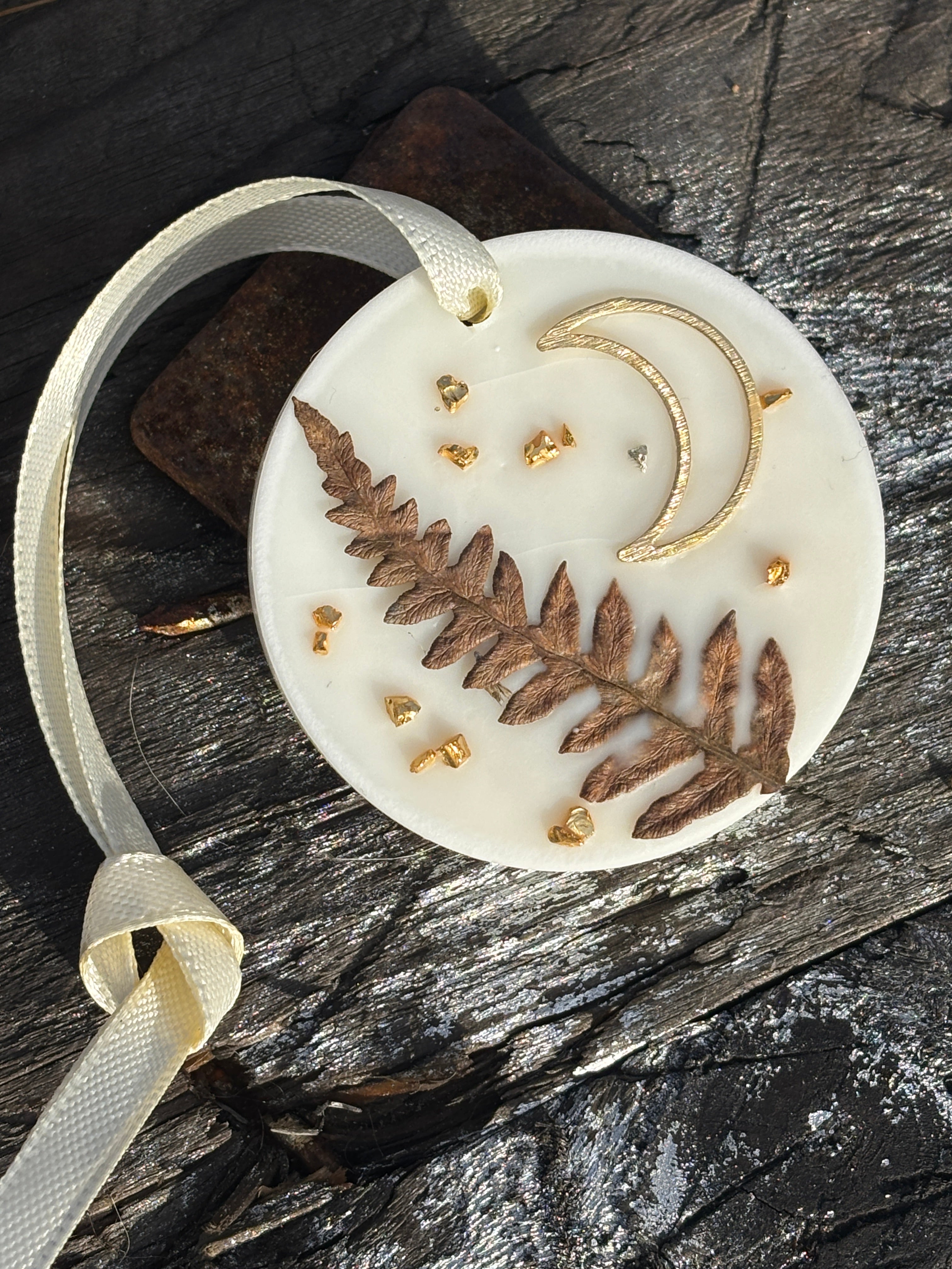 En rund doftvaxhängare som ligger på en träskiva. Hängaren är snygg derokerad med torkade blad, en liten guldmåne och små guldiga glitterbitar.
