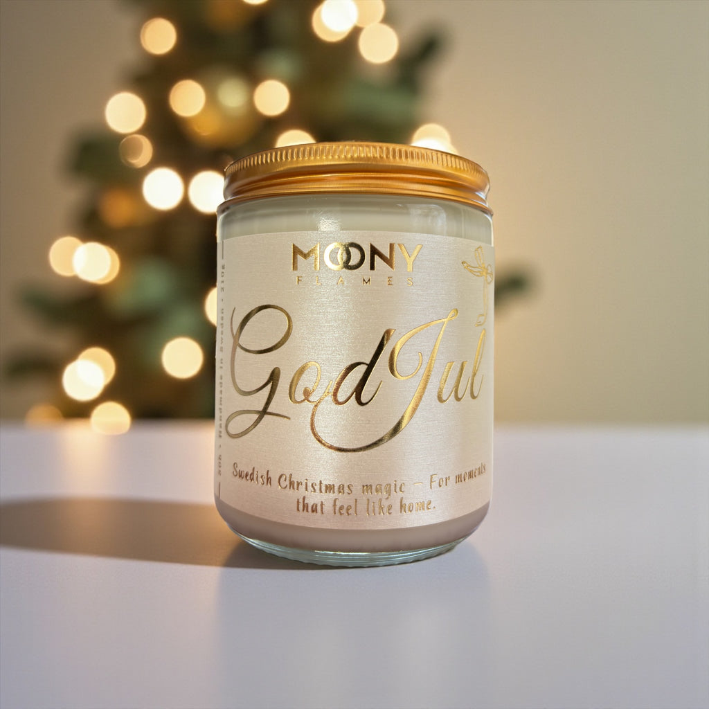 God Jul doftljus som står på ett ljust bord. I bakgrunden syns en tänd julgran.  