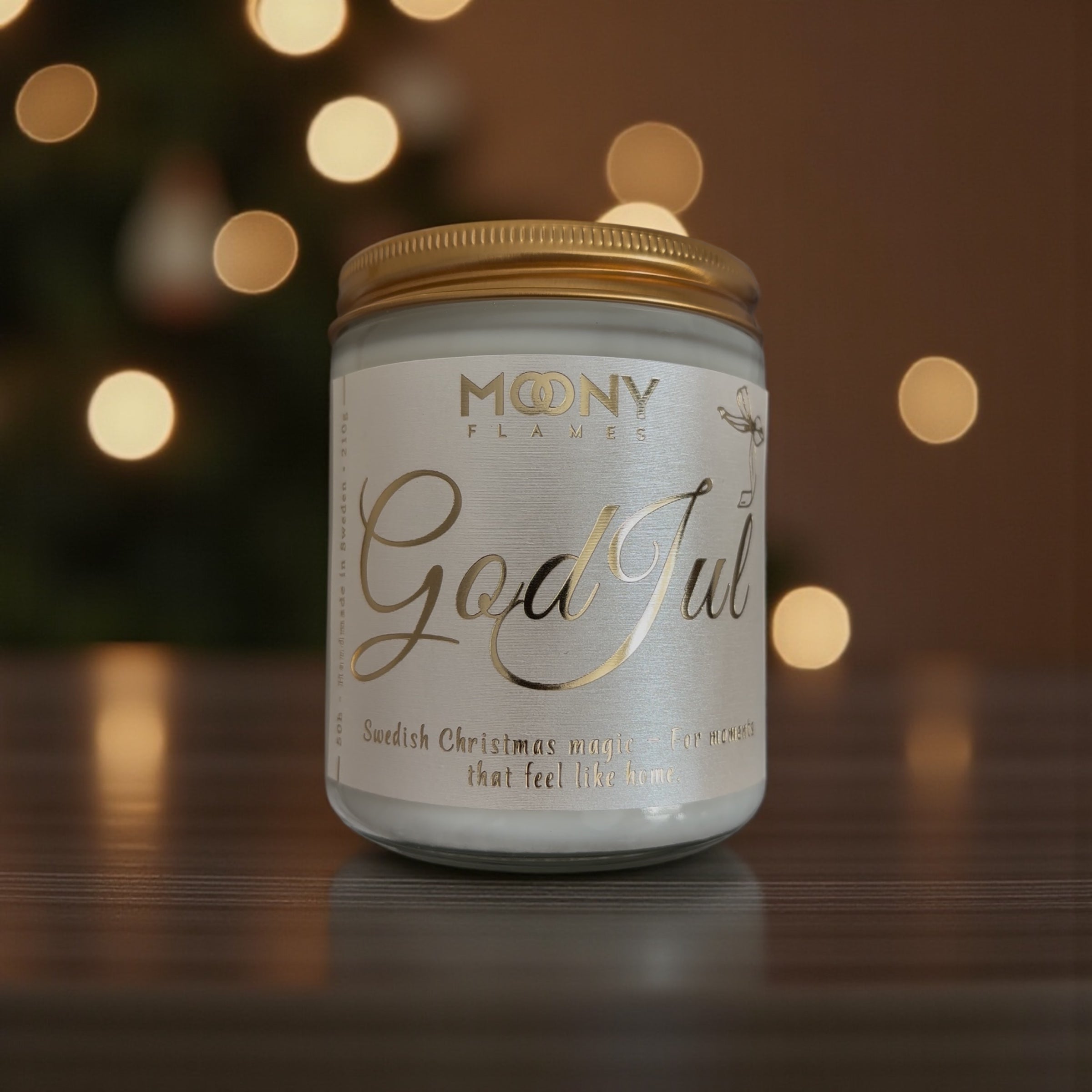 God Jul doftljus som står på ett träbord. I bakrunden syns små lysande ljus från en julgran.