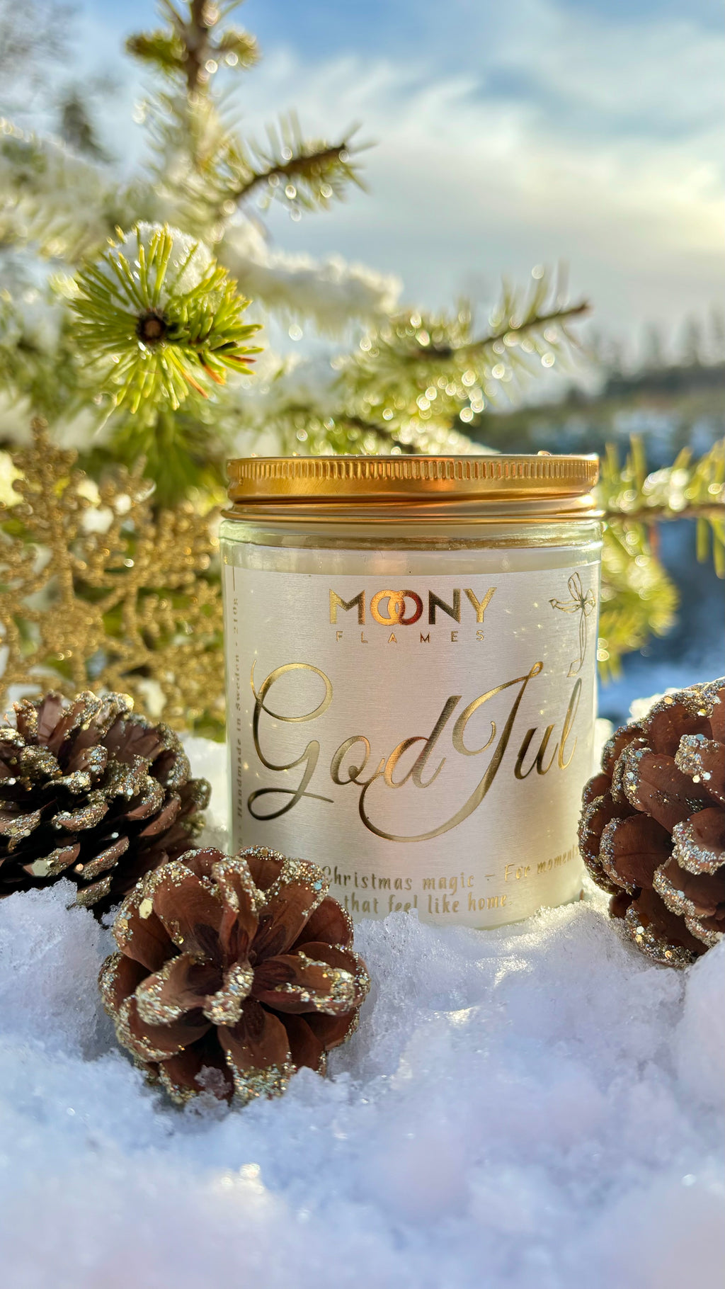 God Jul doftljus som står ute i snön. Runt ljuset syns glittrande grankottar och bakom ljuset ser man en gran.