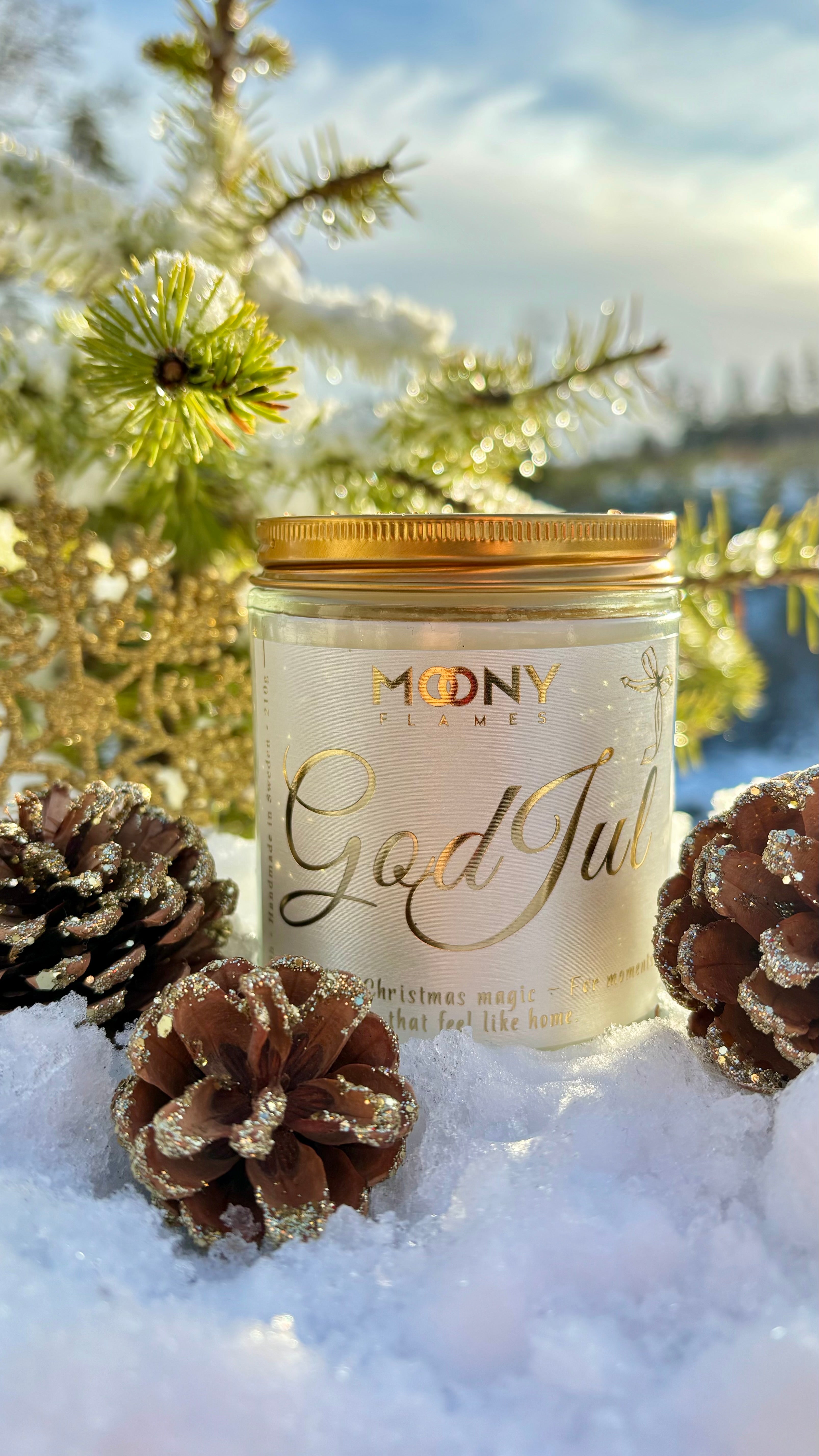 God Jul doftljus som står ute i snön. Runt ljuset syns glittrande grankottar och bakom ljuset ser man en gran.