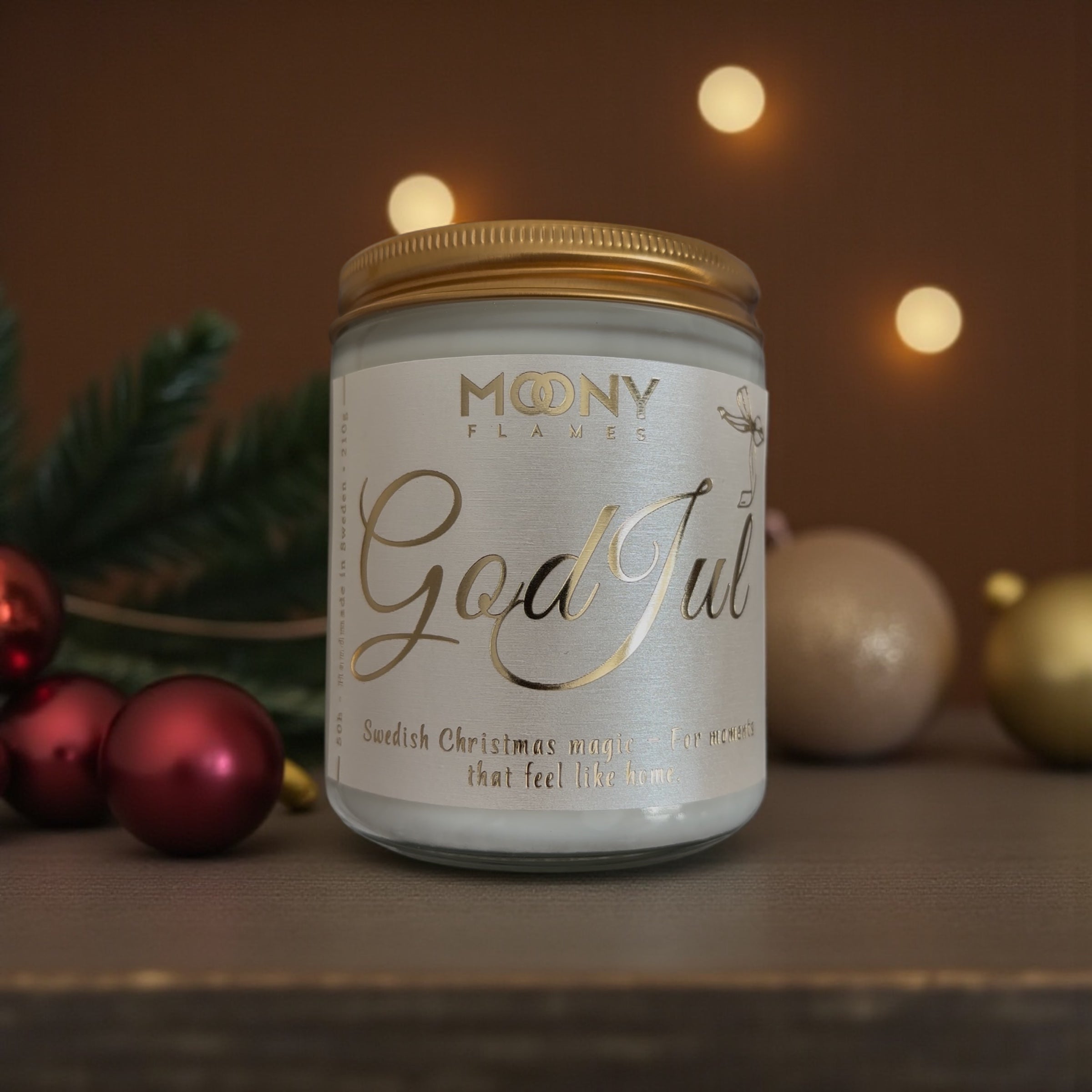 God Jul doftljus som står på ett mörkt bord framför en mörk bakgrund. Runt ljuset ligger röda och glittrande julgranskulor och några gröna kvistar från granen.