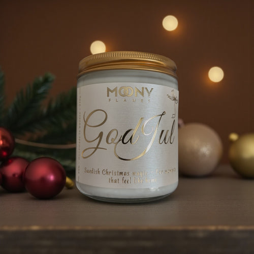 God Jul doftljus som står på ett mörkt bord framför en mörk bakgrund. Runt ljuset ligger röda och glittrande julgranskulor och några gröna kvistar från granen.