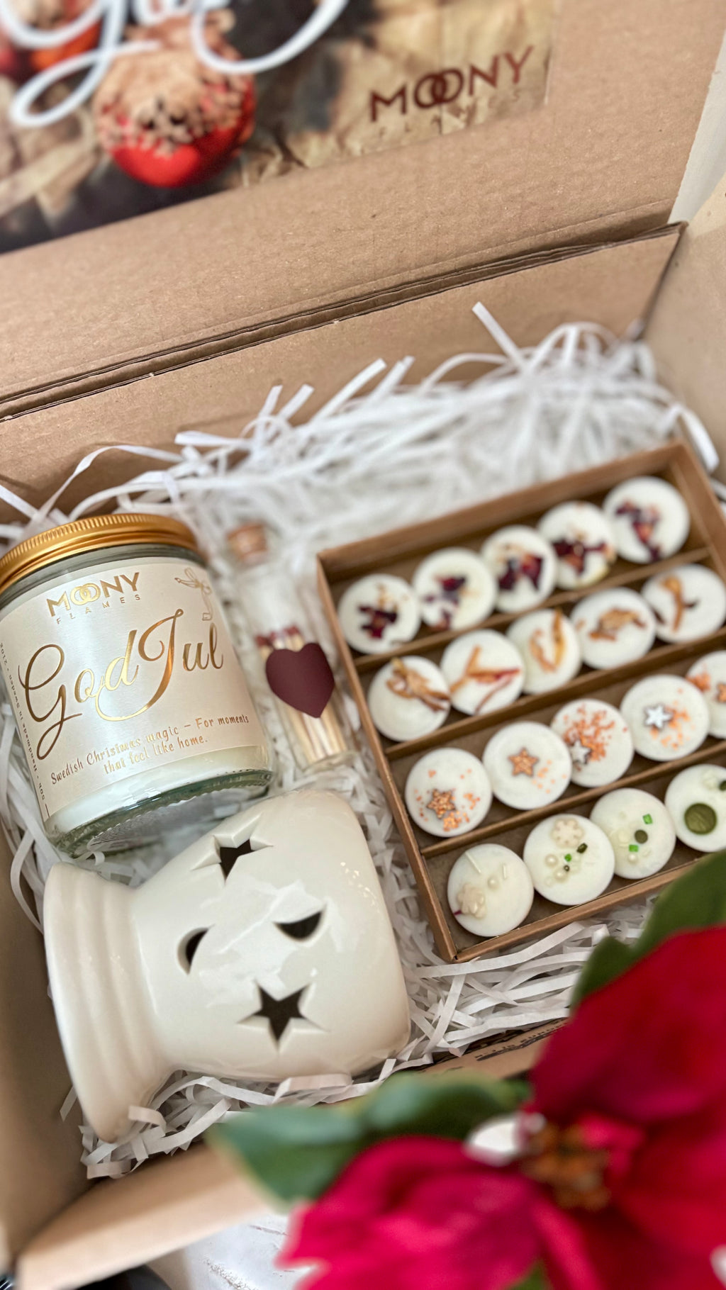 En presentbox som är öppen. Det syns en öppen askmed doftvaxpraliner, Moonys julljus med glänande etikett och en aromalmapa vit. Allting fint dekorerad med juldekorationer.