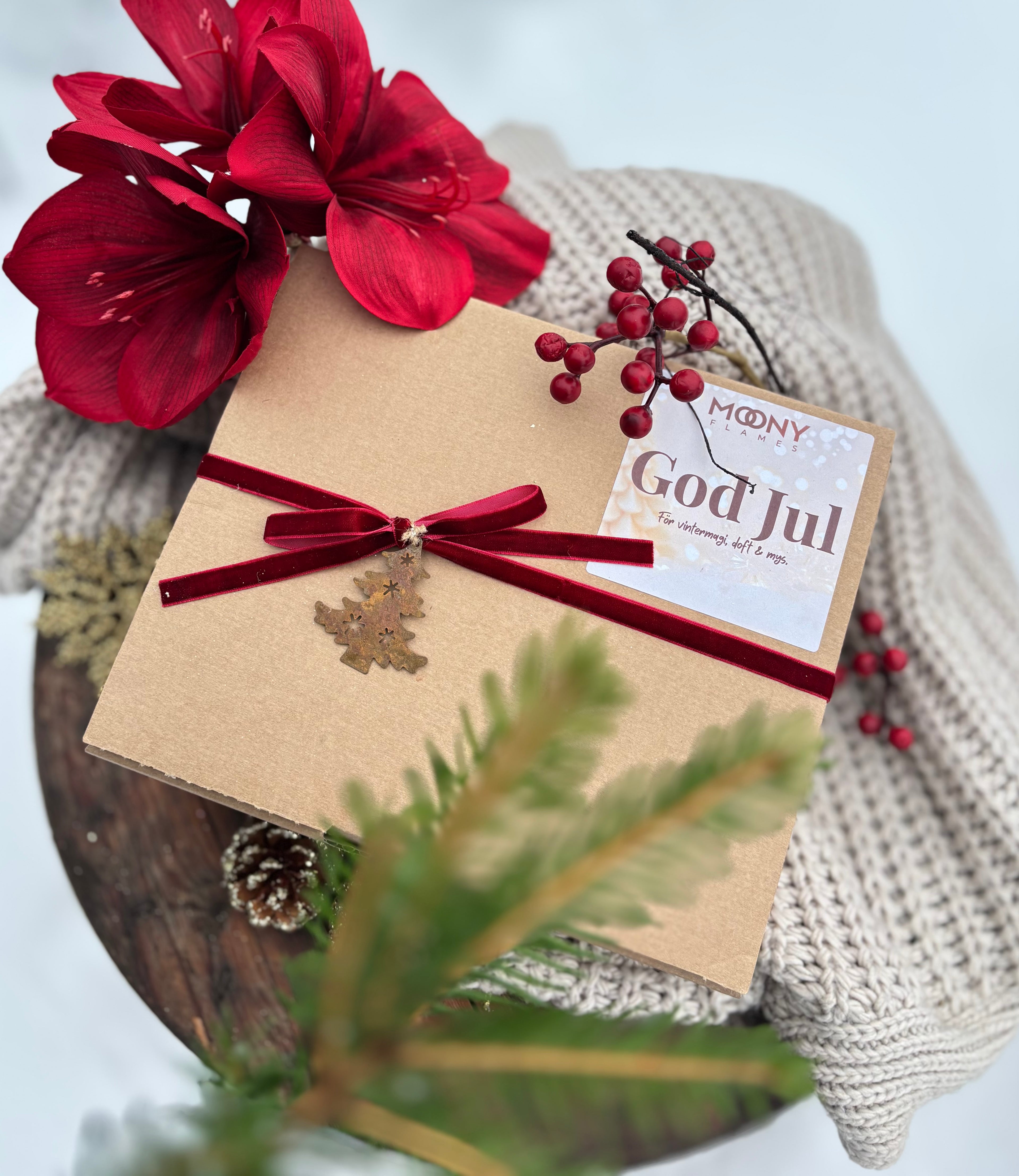 En presentlåda från Moony Flames som ligger på en fild ute i snön. Det syns juliga dekorationer. Lådan försluts med ett rött sammetsband och rosett samt en liten julgransanhängare i rostad metall.