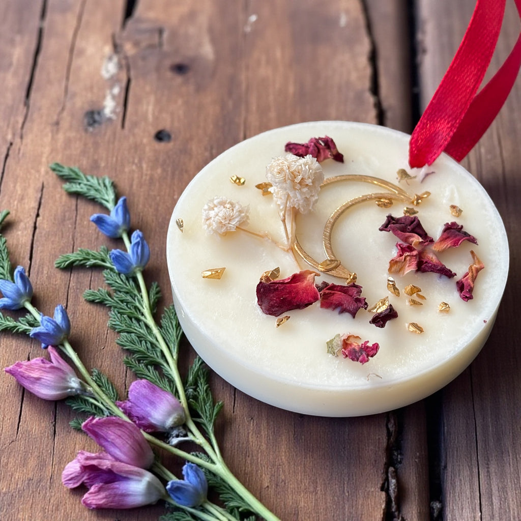 Doftvaxhängare med röd band. Dekorerad med en guldmånde, torkade rosblad som ligger på ett rustikt bord. Det visas några blommor rund hängaren.