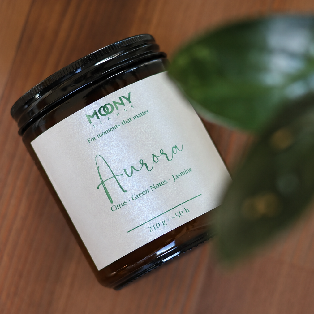 Aurora doftljus från Moony Flames fotograferat ovanifrån på träbord med gröna blad i förgrunden. Handgjort doftljus inspirerat av norrsken med citrus, gröna noter och jasmin, tillverkat i Småland, Sverige.