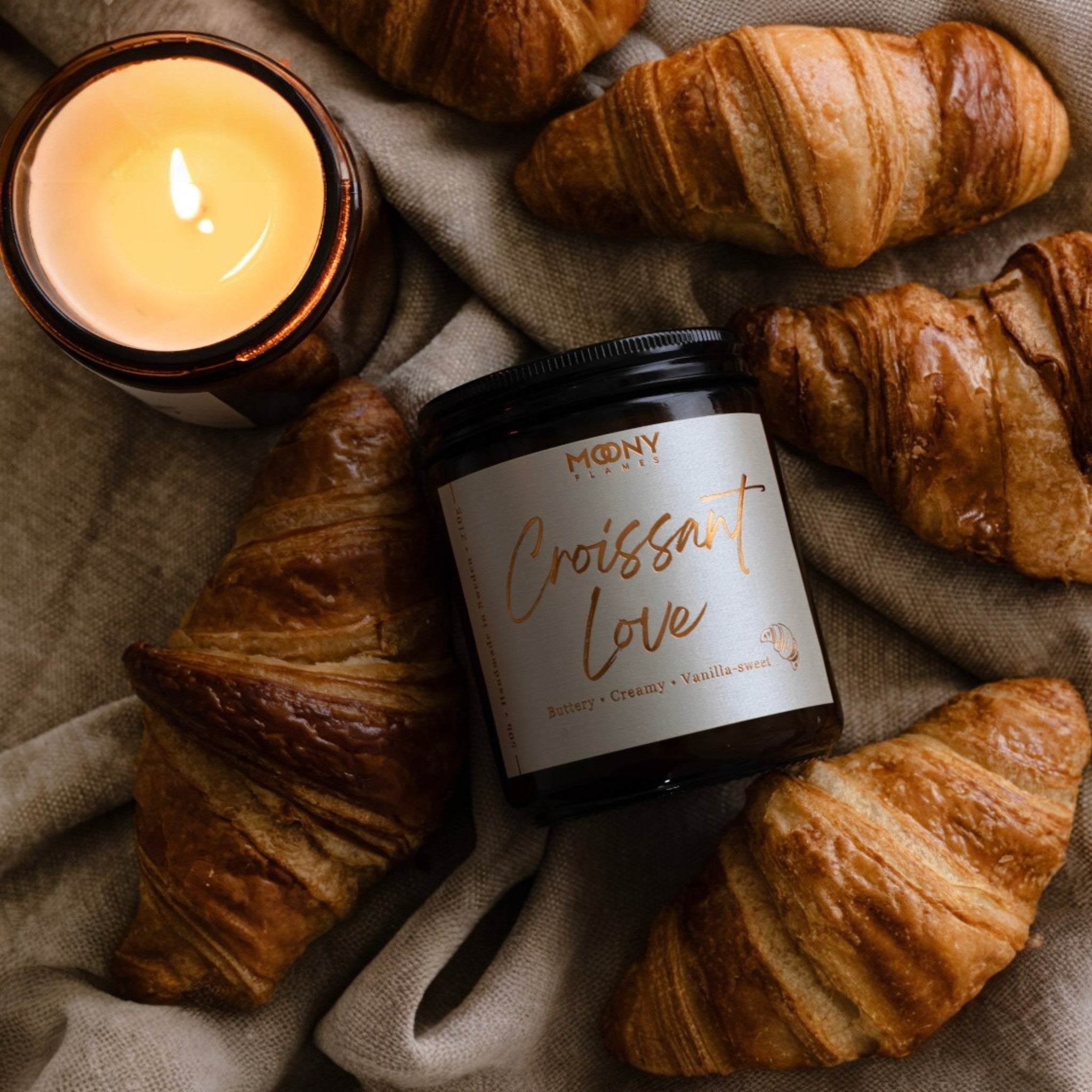 Croissant Love handgjort doftljus från Moony Flames omgivet av nybakade croissanter, inspirerat av känslan av ett franskt bageri en tidig morgon.