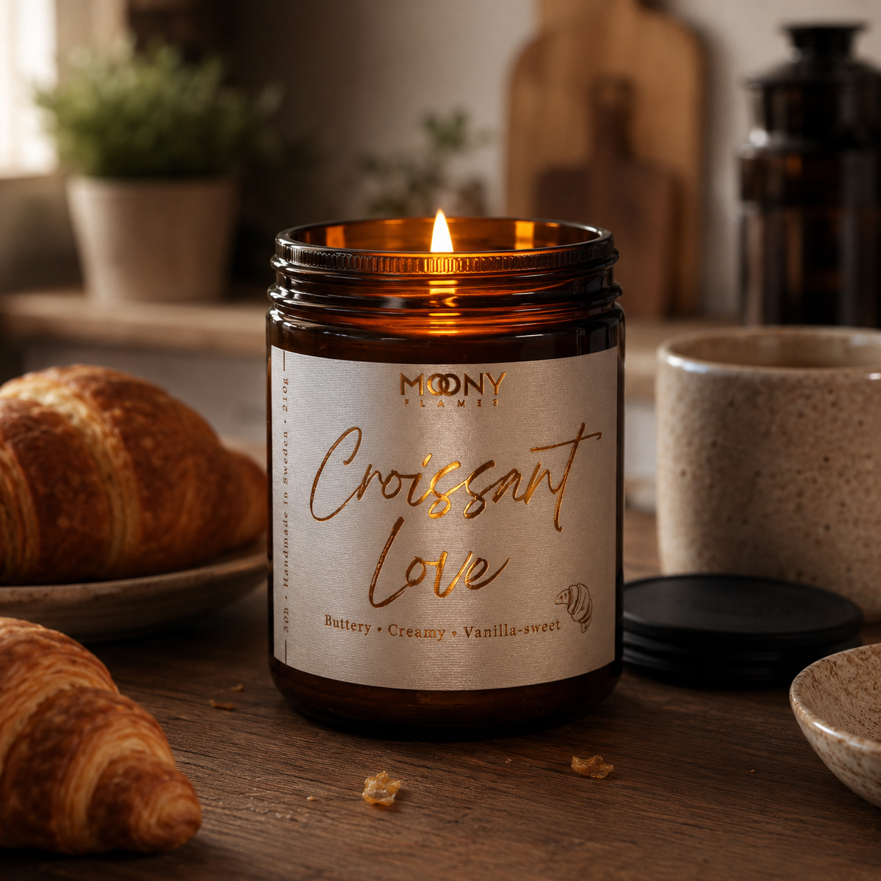 Handgjort Croissant Love doftljus från Moony Flames i amberglas med kopparfolierad etikett. Tänd produktbild i ett mysigt skandinaviskt kök med gyllene croissanter, kaffekopp och rustika trädetaljer. Inspirerat av långsamma helgmorgnar, nybakat franskt bakverk och nybryggt kaffe. Tillverkat i Småland, Sverige av växtbaserat vax.