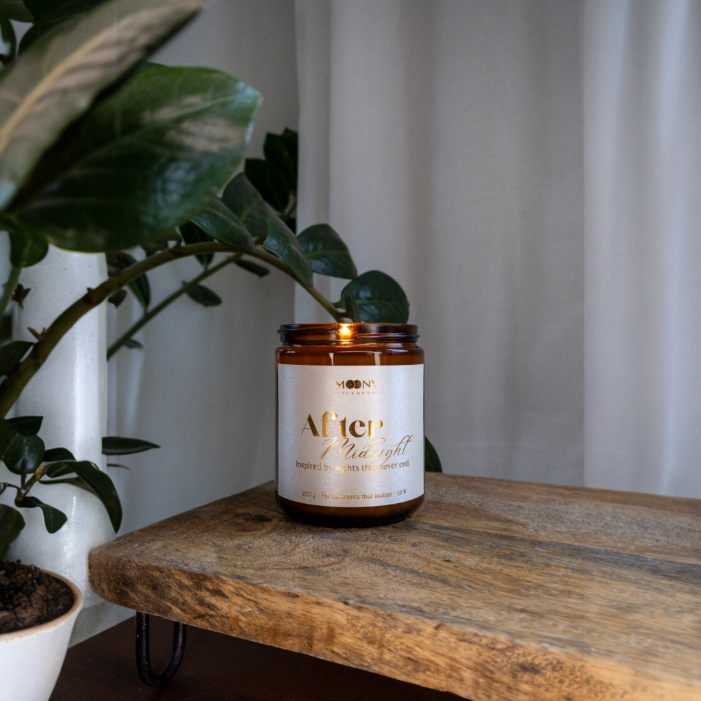 Handgjort doftljus “After Midnight” i amberglas på träbord med gröna växter – lugn och harmonisk hemmiljö