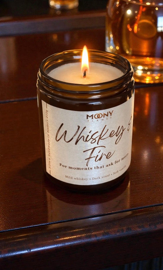 Tänt doftljus Whiskey & Fire från Moony Flames i amberglas, placerat på träyta med varm belysning och stämningsfull hemmiljö i bakgrunden.