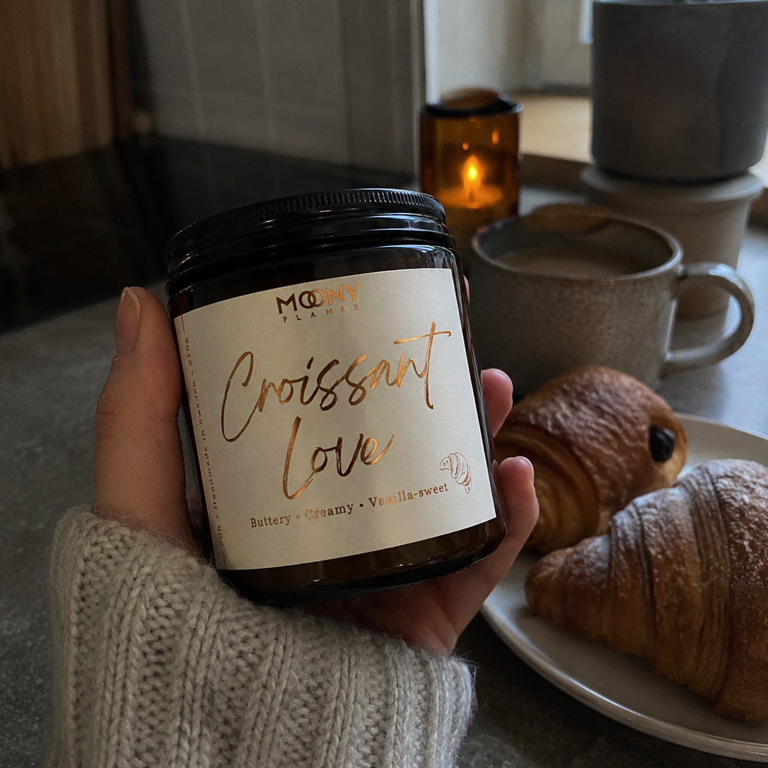 Hand håller handgjorda doftljuset Croissant Love från Moony Flames i ett mysigt kök med nybakade croissanter och kaffe.