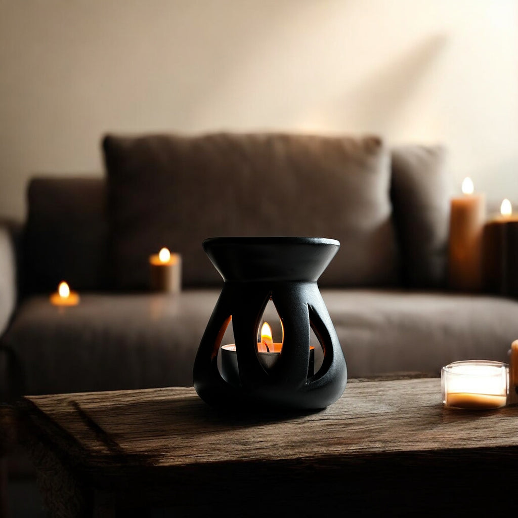 Black Lotus aromalampa i svart keramik – liten doftlampa för doftvax och eteriska oljor som sprider ett mjukt ljus och behaglig doft i ett vardgsrum. Den står på ett träbord och det syns en soffa i bakgrunden samt några tända ljus. 
