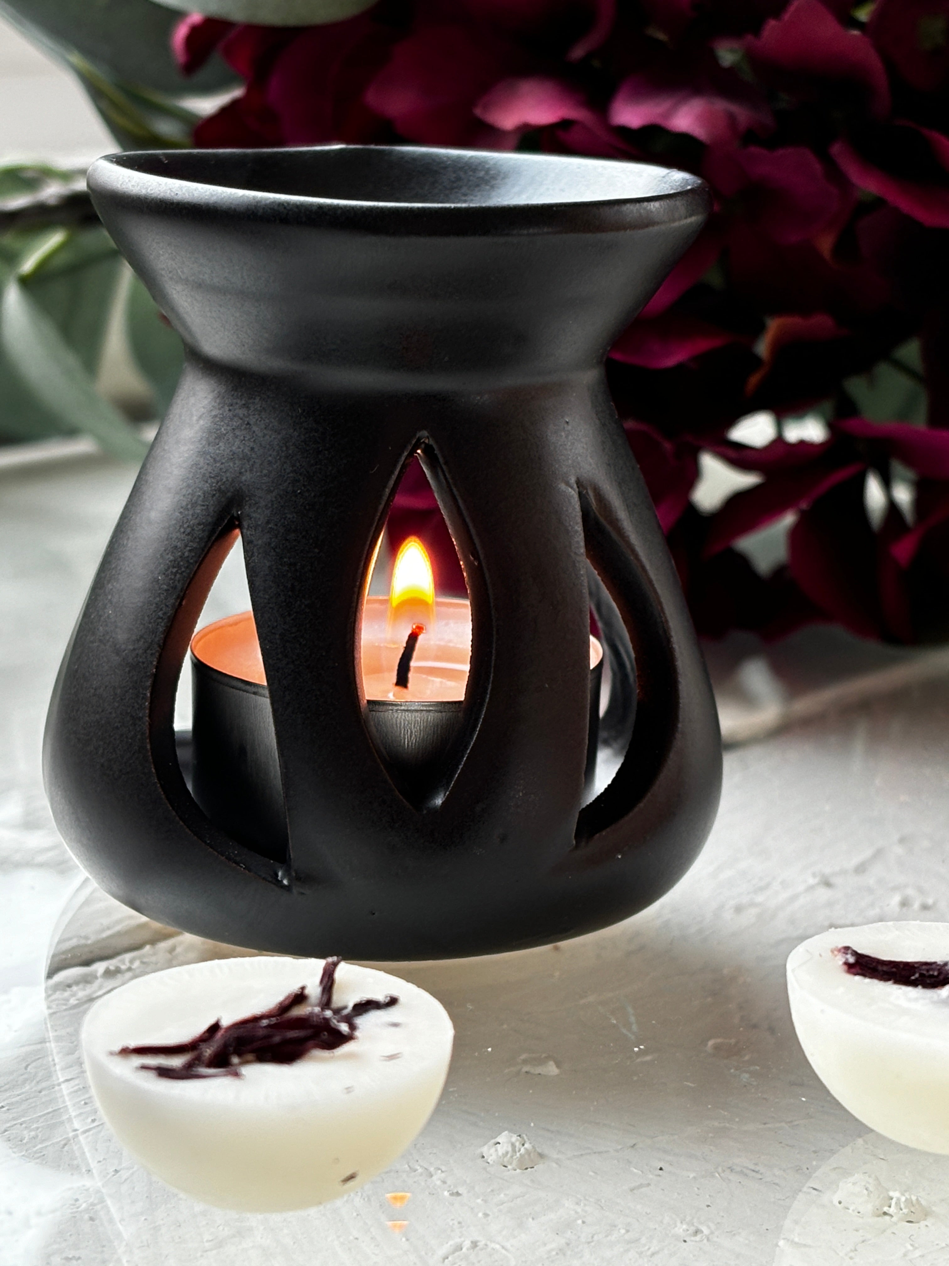 Black Lotus aromalampa i svart keramik – liten doftlampa för doftvax och eteriska oljor som sprider ett mjukt ljus och behaglig doft och som står på ett bord med glasplatta. Det visas några doftpraliner framför ljuset samt torkade blommer bakom ljuset. Lampan är tänd..