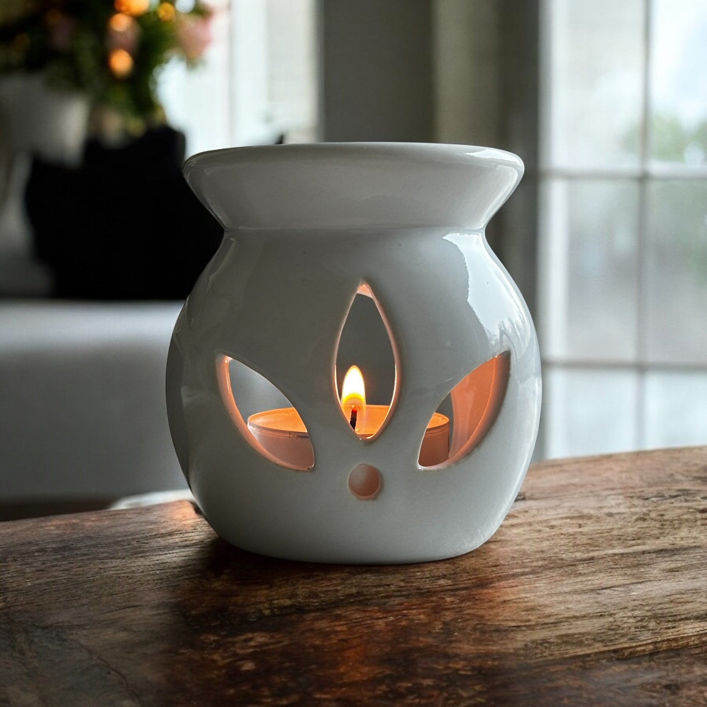 White Lotus aromalampa i vit keramik – liten och elegant doftlampa för doftvax och eteriska oljor som är tänd och som står på en rustik hylla. I bakgrunden syns ett fönster och en soffa.
