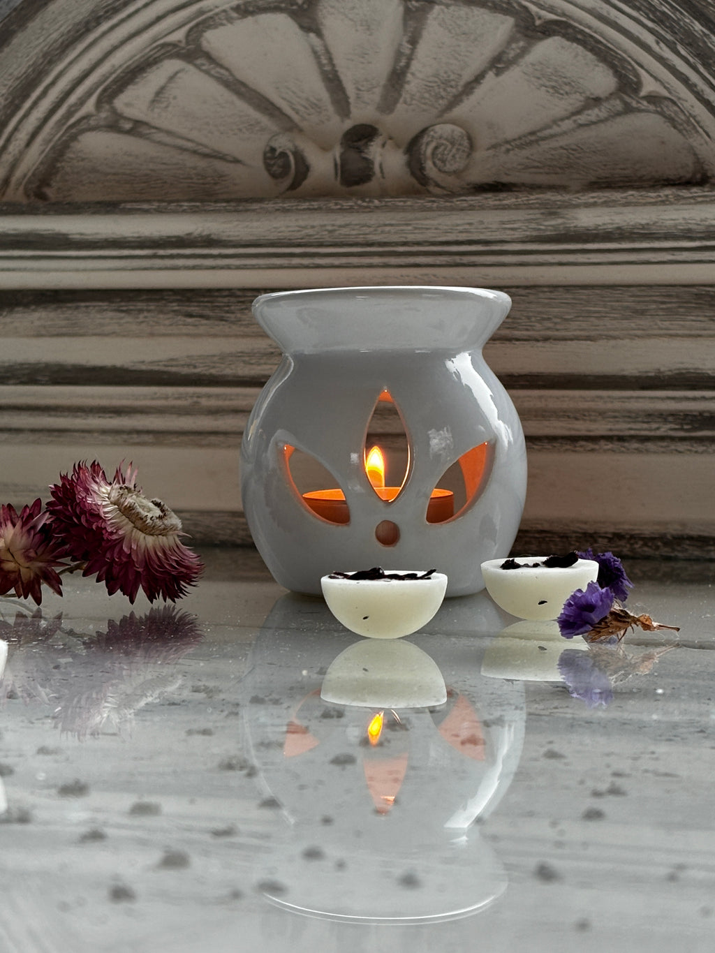 White Lotus aromalampa i vit keramik – liten och elegant doftlampa för doftvax och eteriska oljor som står på ett glasbord och det ligger några doftpraliner och torkade blomor framför den. 