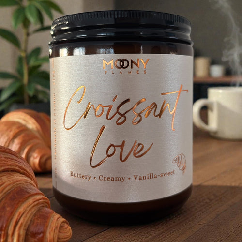 Handgjutet doftljus Croissant Love från Moony Flames i bärnstensfärgad glasburk med svart lock och krämfärgad etikett med kopparfolie, placerat på träbord bredvid nybakade croissanter i varm köksmiljö.
