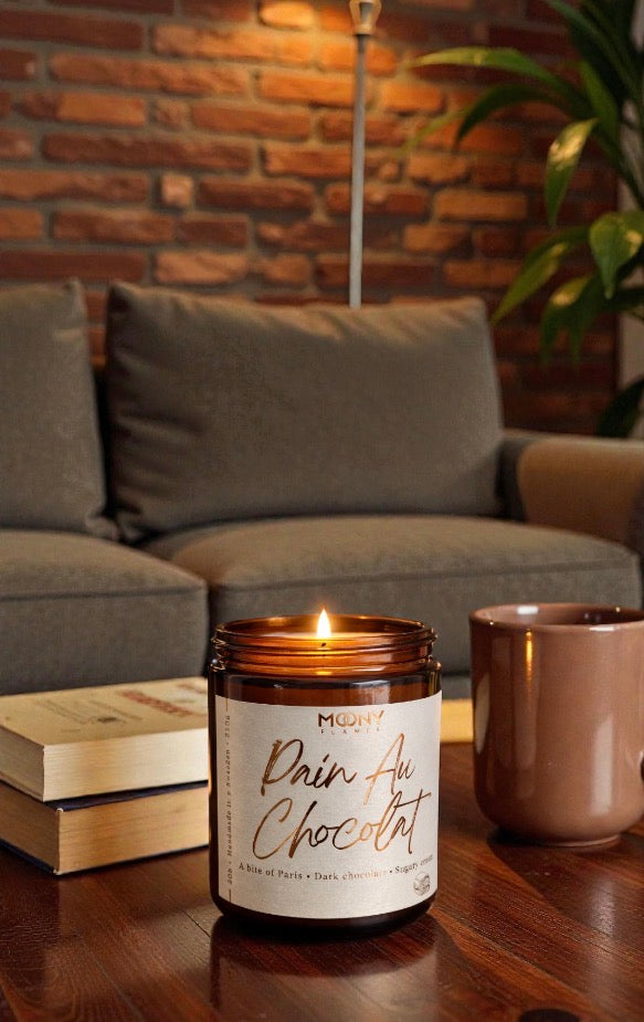 Brinnande Pain Au Chocolat doftljus från Moony Flames i vardagsrumsmiljö, med doft av mörk choklad och söt grädde, placerat vid bok och kaffekopp för en varm och inbjudande stund.