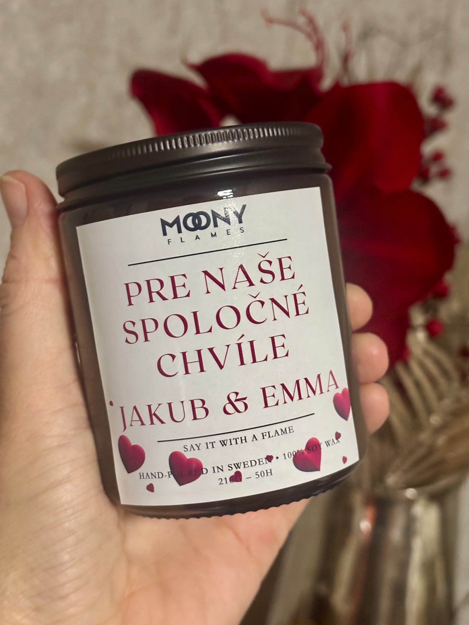 Handgjutet personligt doftljus från Moony Flames med texten ”Pre naše spoločné chvíle – Jakub & Emma”, hålls i handen framför röda blommor i romantisk miljö.