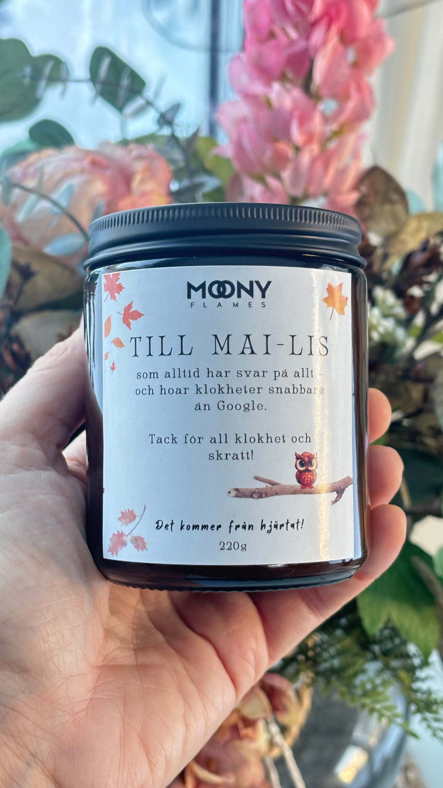 Handgjutet personligt doftljus från Moony Flames med personlig hälsning till ”Till Mai-Lis”, hålls i handen framför blommor i ljus hemmiljö.