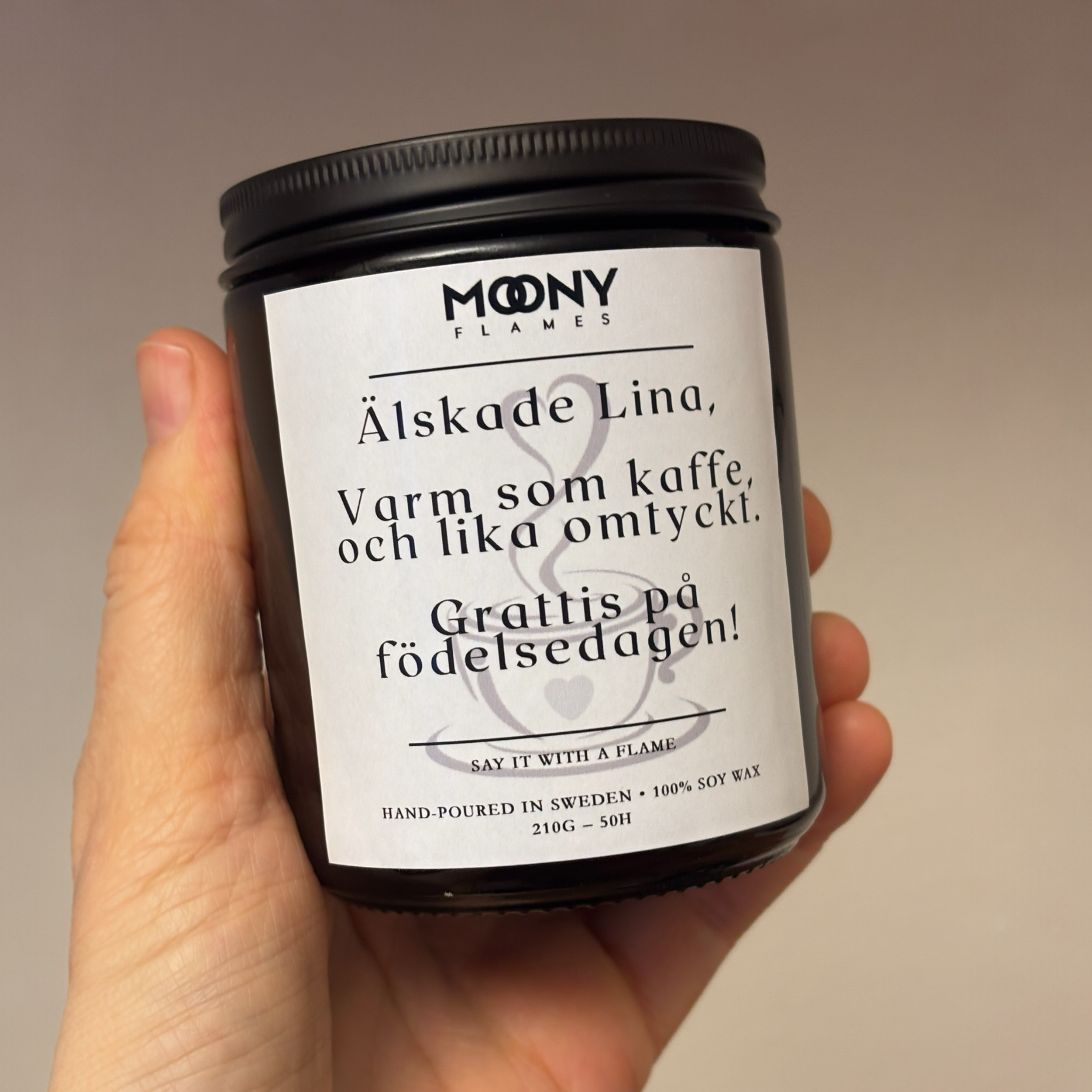 Ett doftljus med budskap och egen text på etikett från Moony Flames Sverige som hålls i handen framför en varm beige bakgrund.