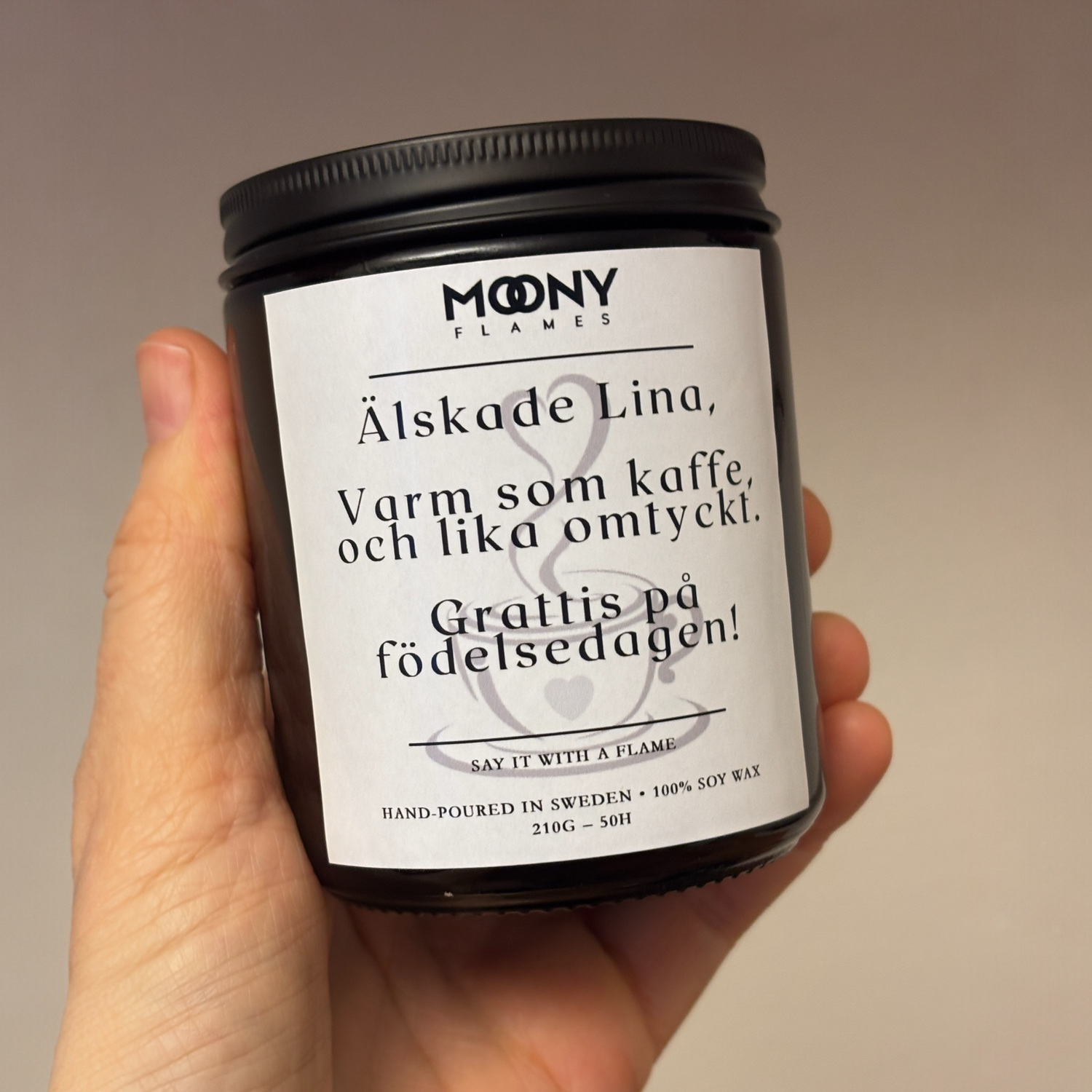 Ett doftljus med budskap och egen text på etikett från Moony Flames Sverige som hålls i handen framför en varm beige bakgrund.
