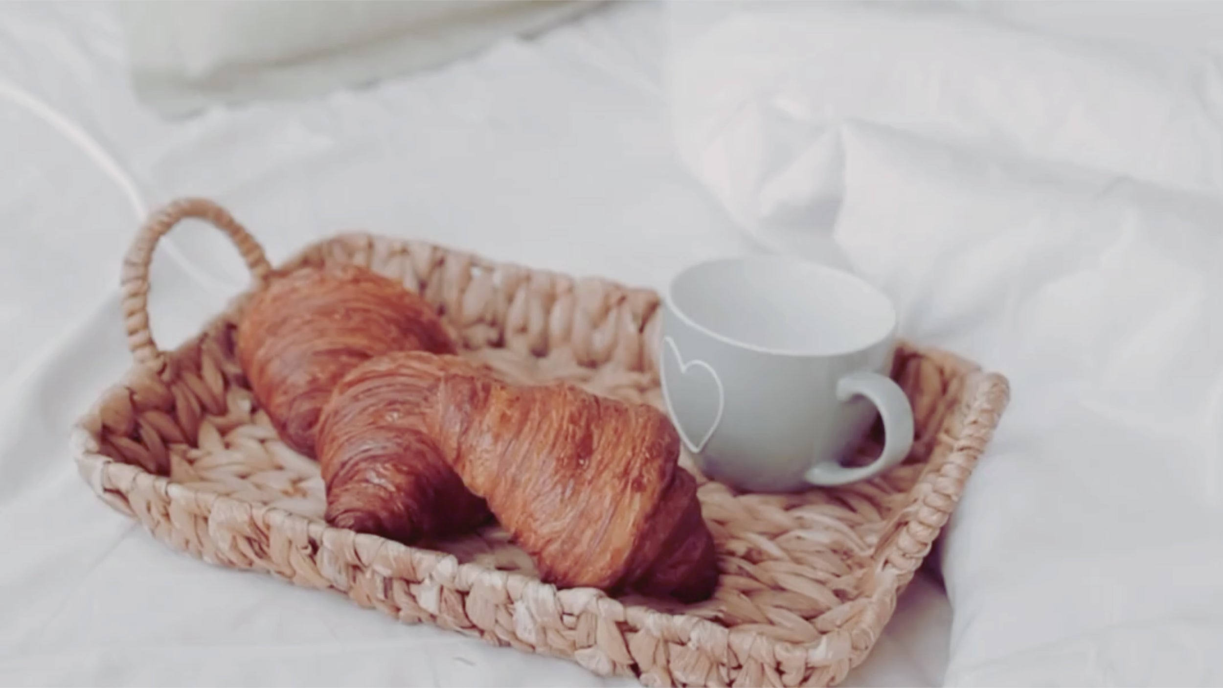I videon visas en brödkorg med nybakade croissanter och en kaffekopp som tår på en mysig säng i morgonljuset.