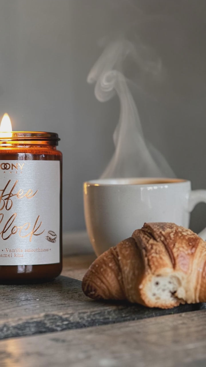 Handgjort doftljus Coffee O’Clock i brun glasburk bredvid en rykande kopp kaffe och croissant – varm, mysig morgonstämning.