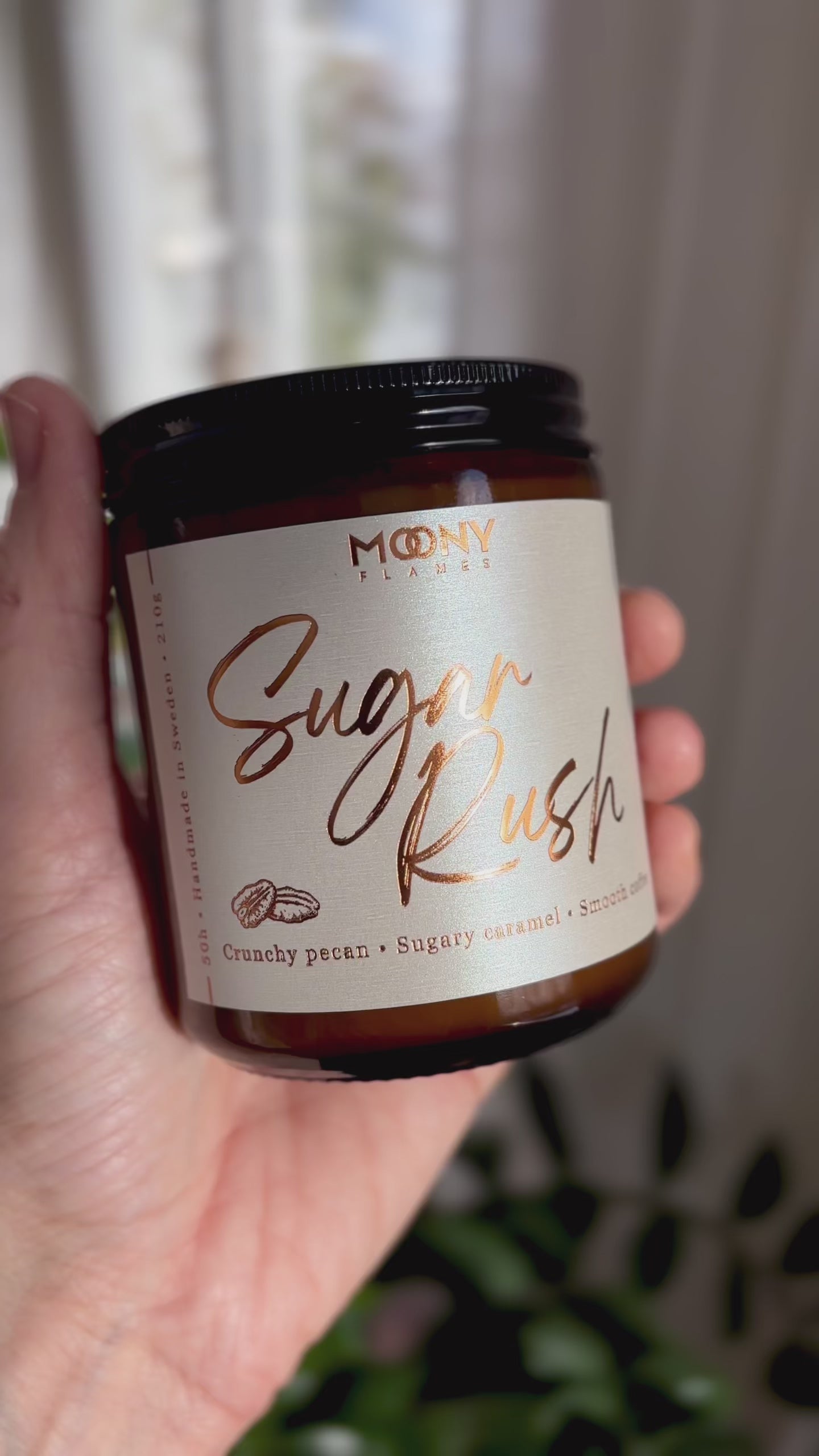 Hand som håller Sugar Rush doftljus från Moony Flames i amberglas, handgjutet doftljus med karamell- och pekannötstoner.