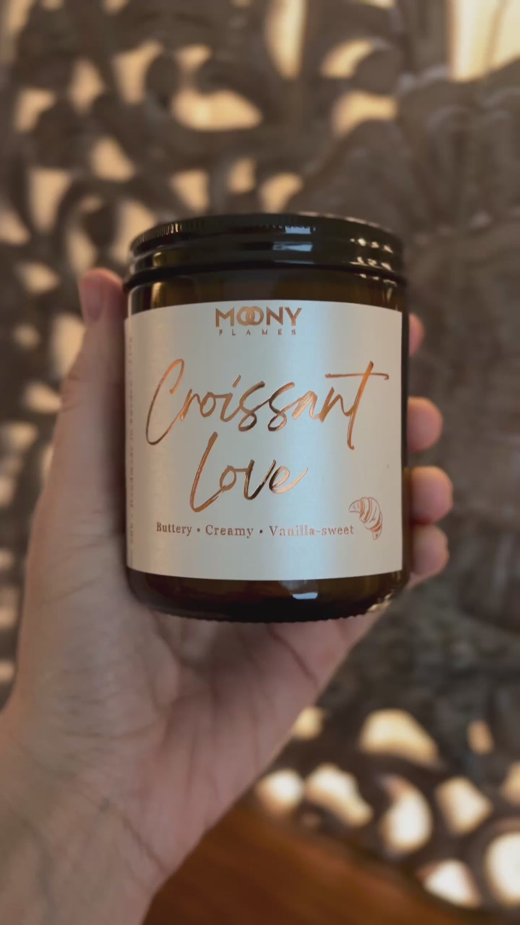 Handhållet doftljus från Moony Flames med namnet Croissant Love i bärnstensfärgad glasburk. Etiketten är krämvit med kopparfärgad text och beskriver doften som smörig, krämig och vaniljsöt.