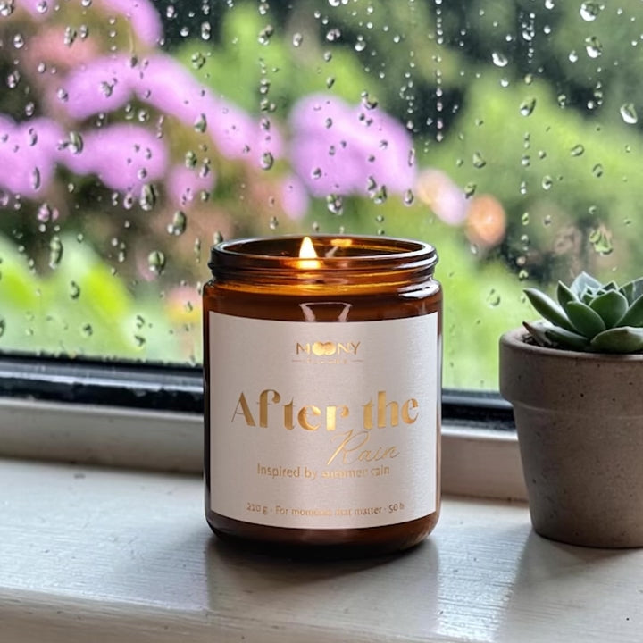 Handgjort doftljus After the Rain som brinner framför ett fönster med regn utanför – inspirerat av sommarregn och petrichor.